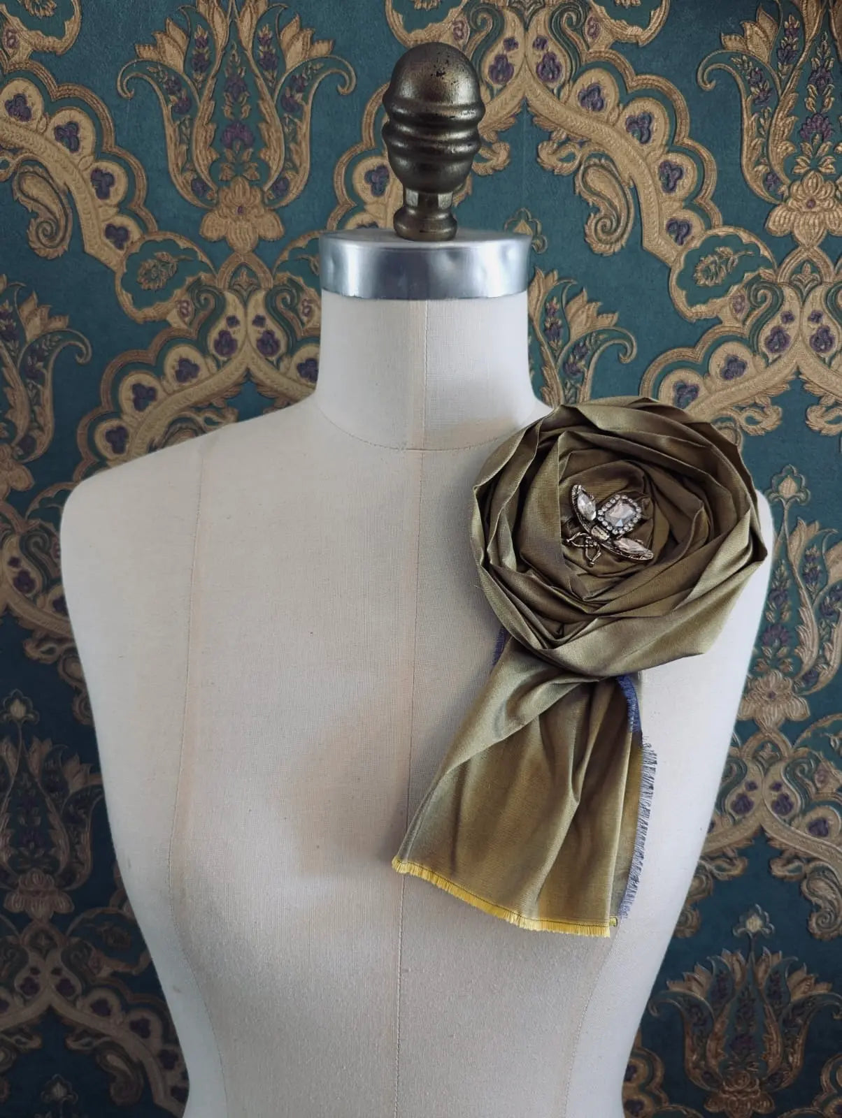 Broochella rosa ingioiellata gold handmade designer silk silk flower brooch with rhinestone insect embellishment. styled on a mannequin. The background is green and gold damask wallpaper