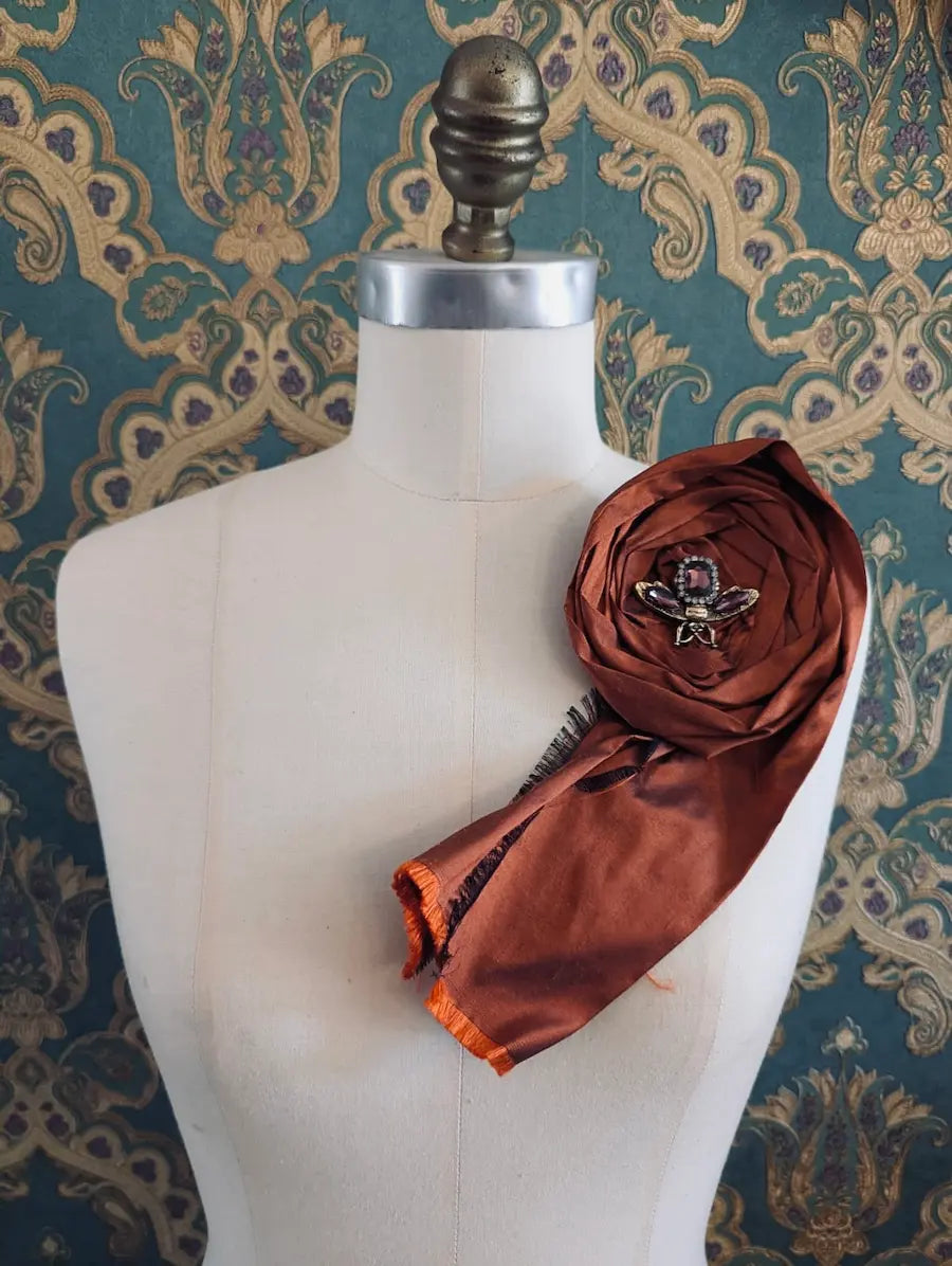 Broochella rosa ingioiellata rust handmade designer silk silk flower brooch with rhinestone insect embellishment. styled on a mannequin. The background is green and gold damask wallpaper