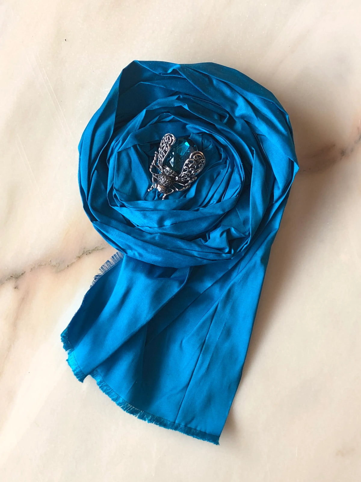 Broochella Rosa Ingioiellata Large handmade designer flower brooch in vivid blue silk, sculpted into an oversized rose and finished with a tonal blue crystal jewelled insect centrepiece, photographed on cream marble with natural grey veining.