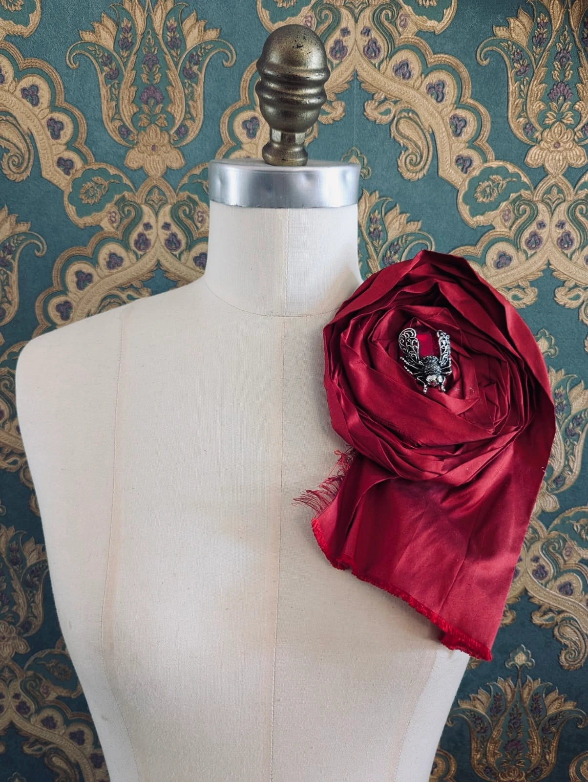 Broochella rosa ingioiellata large red handmade silk flower brooch with an insect brooch embellishment in the centre. It is pinned to a mannequin. The background is green and gold damask wallpaper.