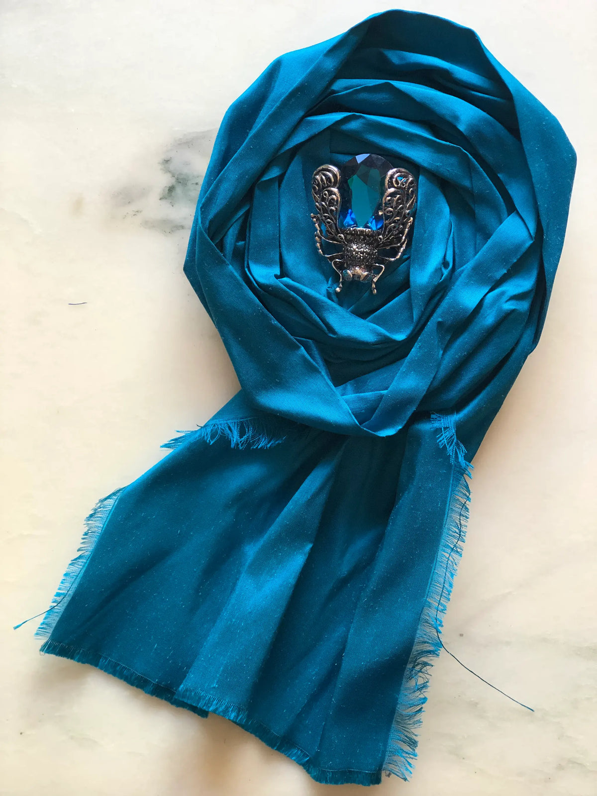Broochella Rosa Ingioiellata Flower Brooch Pin - Small in rich teal blue silk, styled with a vintage blue crystal and silver-tone brooch at its centre on a cream marble background.