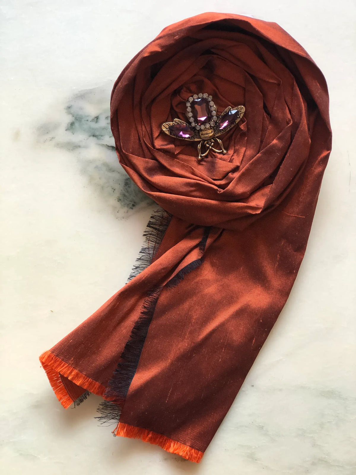 Broochella Rosa Ingioiellata Flower Brooch Pin - Small in rich rust silk, styled with a vintage blue crystal and silver-tone brooch at its centre on a cream marble background.