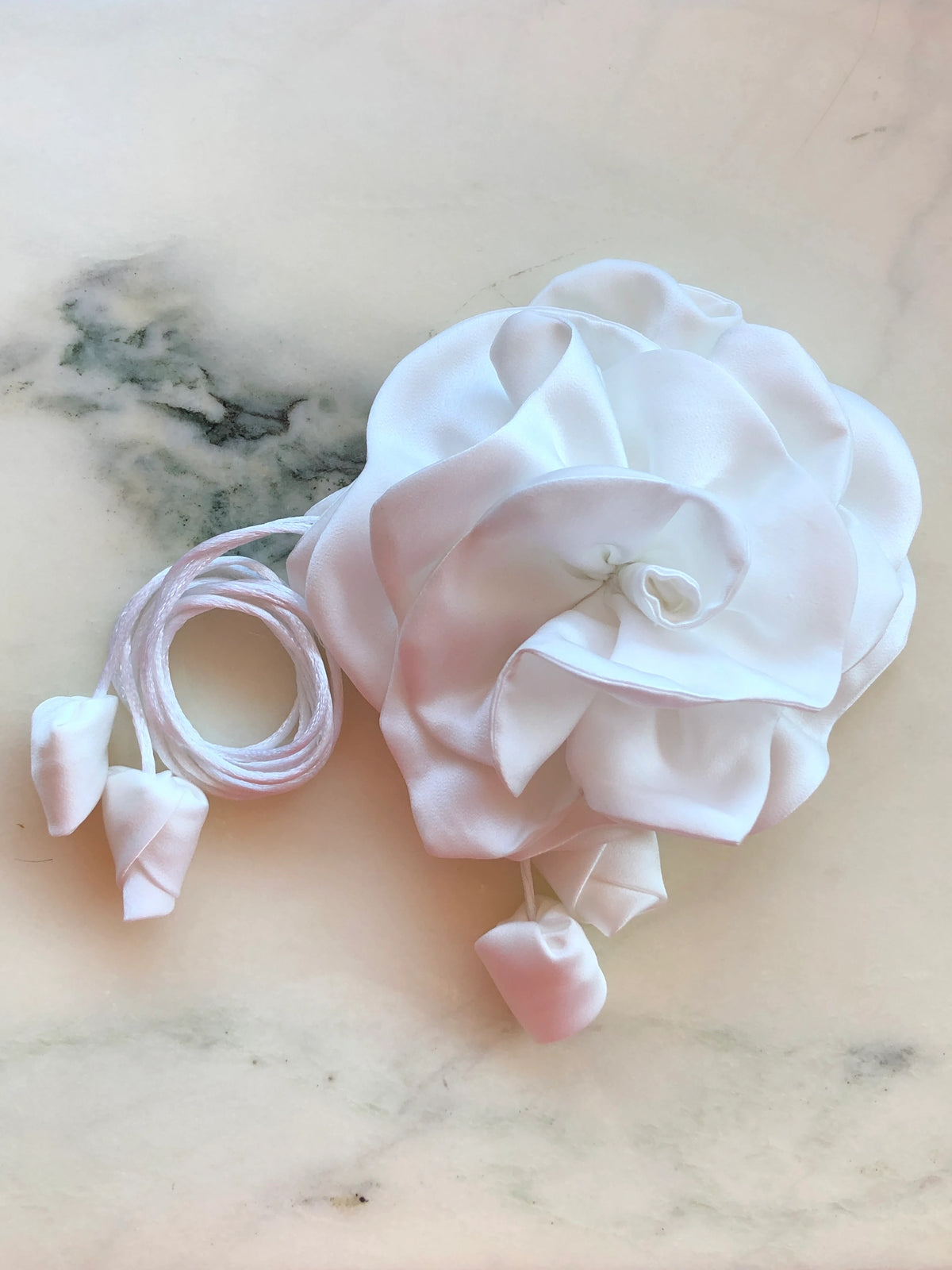 Broochella Rosetta Bridal Choker / Brooch in white, featuring an oversized sculptural satin rose with coordinating satin ties and petal-shaped ends, photographed on a pale cream marble background.