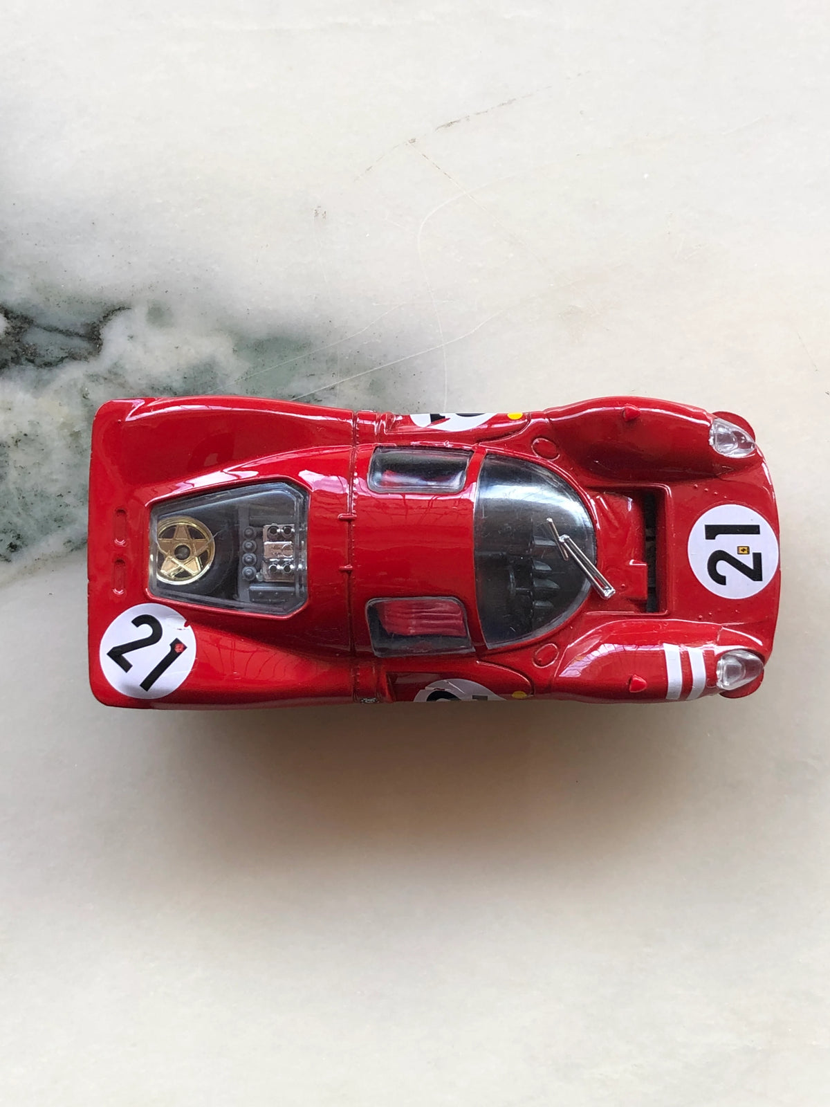 Broochella Rosso Fiorentino No.21 Men’s Racing Car Brooch photographed from above, showcasing its red aerodynamic body, white number 21 racing decals, transparent cockpit canopy and detailed rear engine compartment.