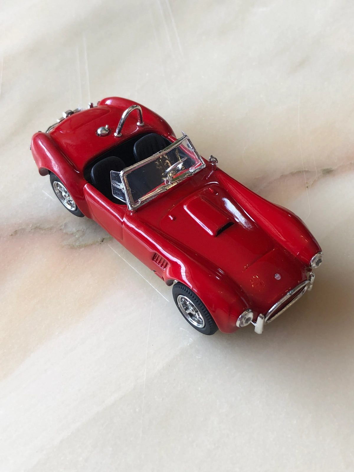 Broochella Rosso Roadster No.427 Vintage Convertible Racing Car Brooch in glossy red, featuring black seats, a clear windscreen, chrome roll bar, silver wire-spoke wheels and classic roadster detailing, photographed from an elevated angle.