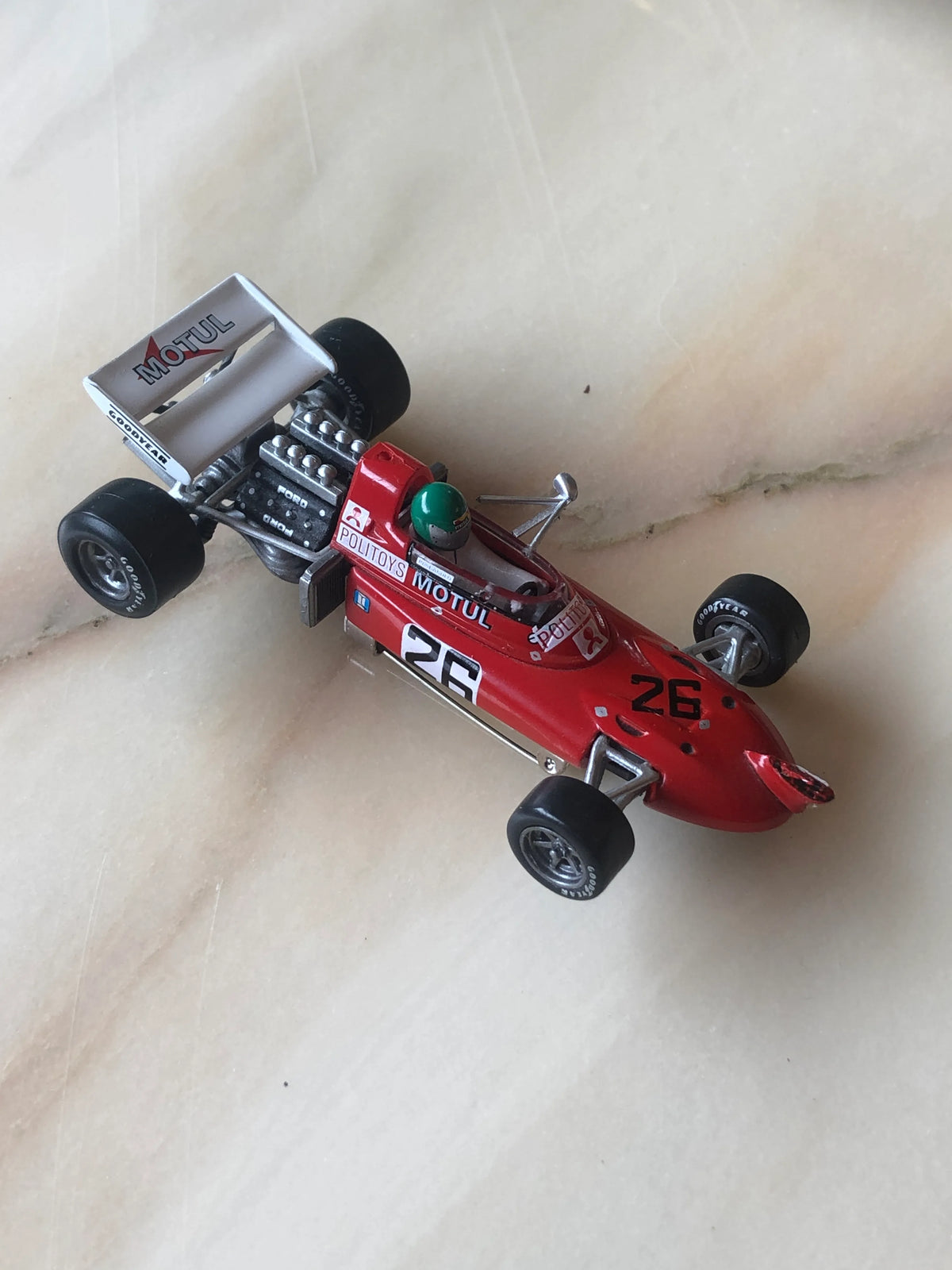 Broochella Rosso Turbo No.26 Vintage Formula Racing Car Brooch photographed at an angle, showcasing its bright red aerodynamic body, number 26 racing decals, green helmet, detailed exposed engine, black Goodyear tyres and white rear spoiler.