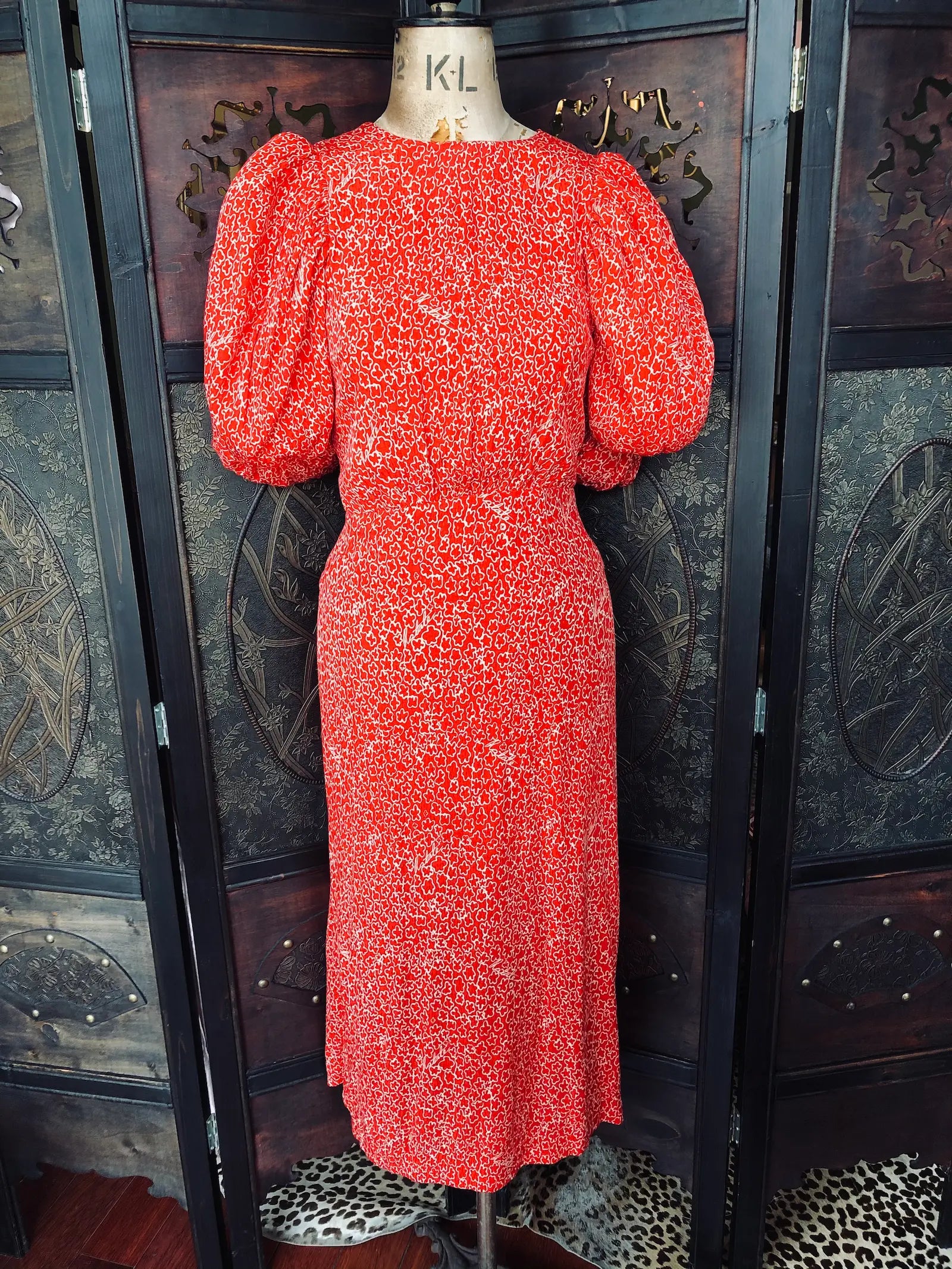 Broochella Rotate red and white patterned midi dress with puffed short sleeves is displayed on a vintage mannequin torso in front of a dark wooden folding screen.