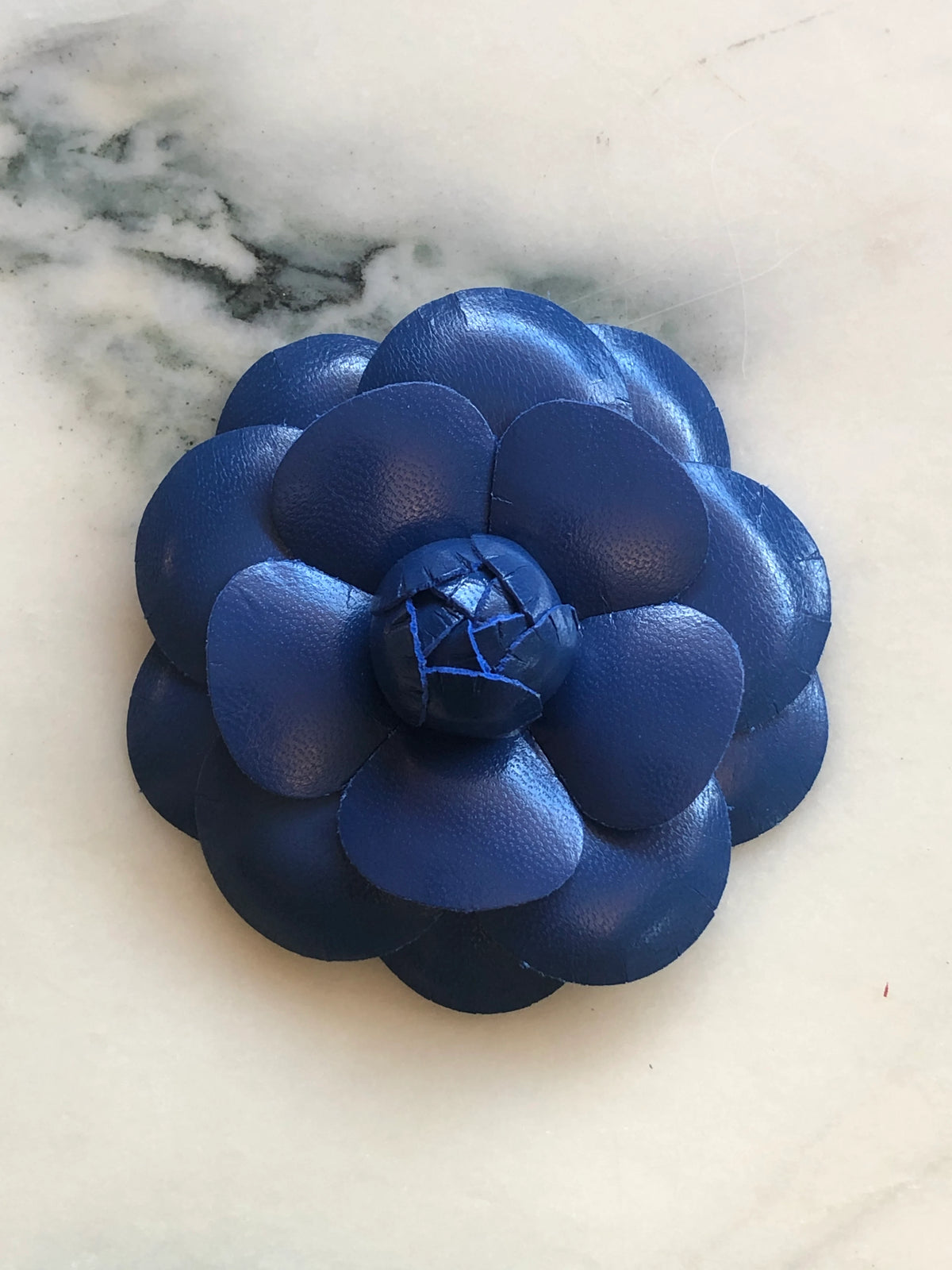 A Broochella Camelia handmade royal blue leather flower-shaped accessory brooch,  resting on a white marble surface.