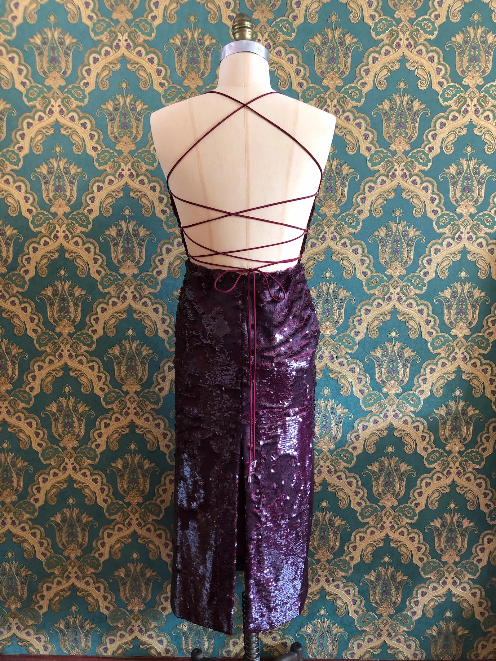 The back view of Broochella Sabina Musáyev Zarya midi dress in Ruby Wine with tonal sequins, cowl neckline and lace-up back, size XS. Pictures on a mannequin. The background is green and gold damask wallpaper