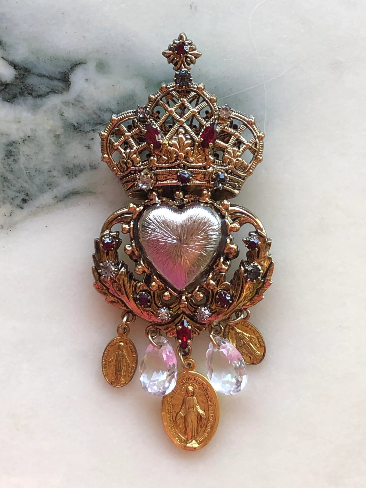 Broochella Sacro Cuore Agatha Brooch in ornate gold with a red heart, jewelled crown, crystal drops and devotional medallions, photographed on a white marble background.