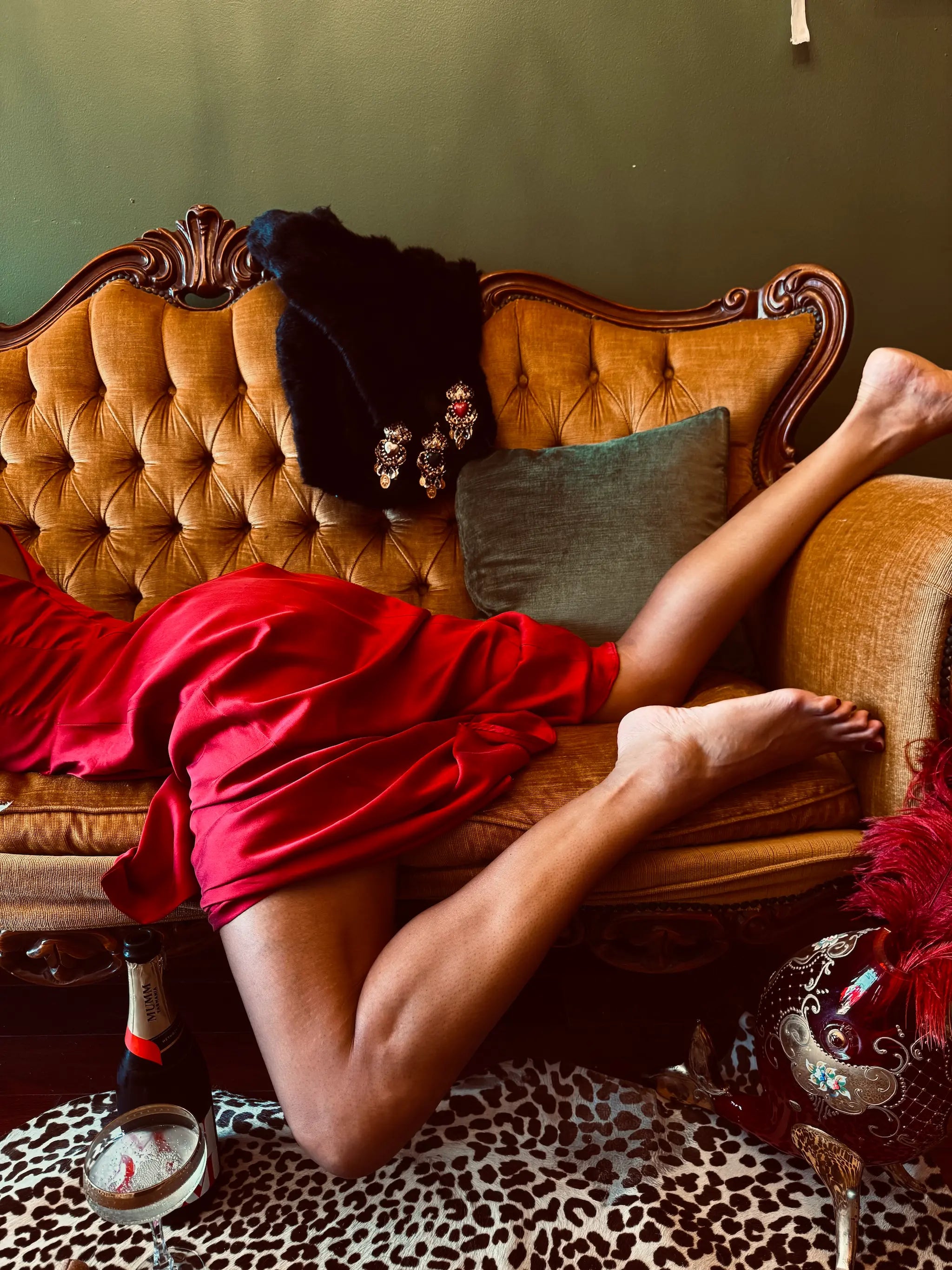 Broochella agatha brooches are pinned to a black coat and draped over a gold couch with a wooden frame. A model is laying on her stomach on a red dress. Her legs are stretched across the couch.
