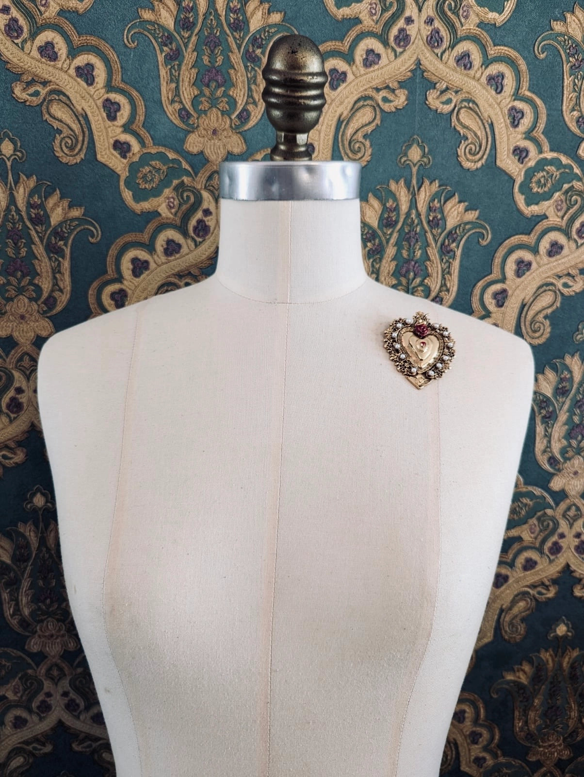 Broochella Sacro cuore benedetta black and gold metal  heart shaped brooch. Pinned to a mannequin. The background is green and gold damask wallpaper