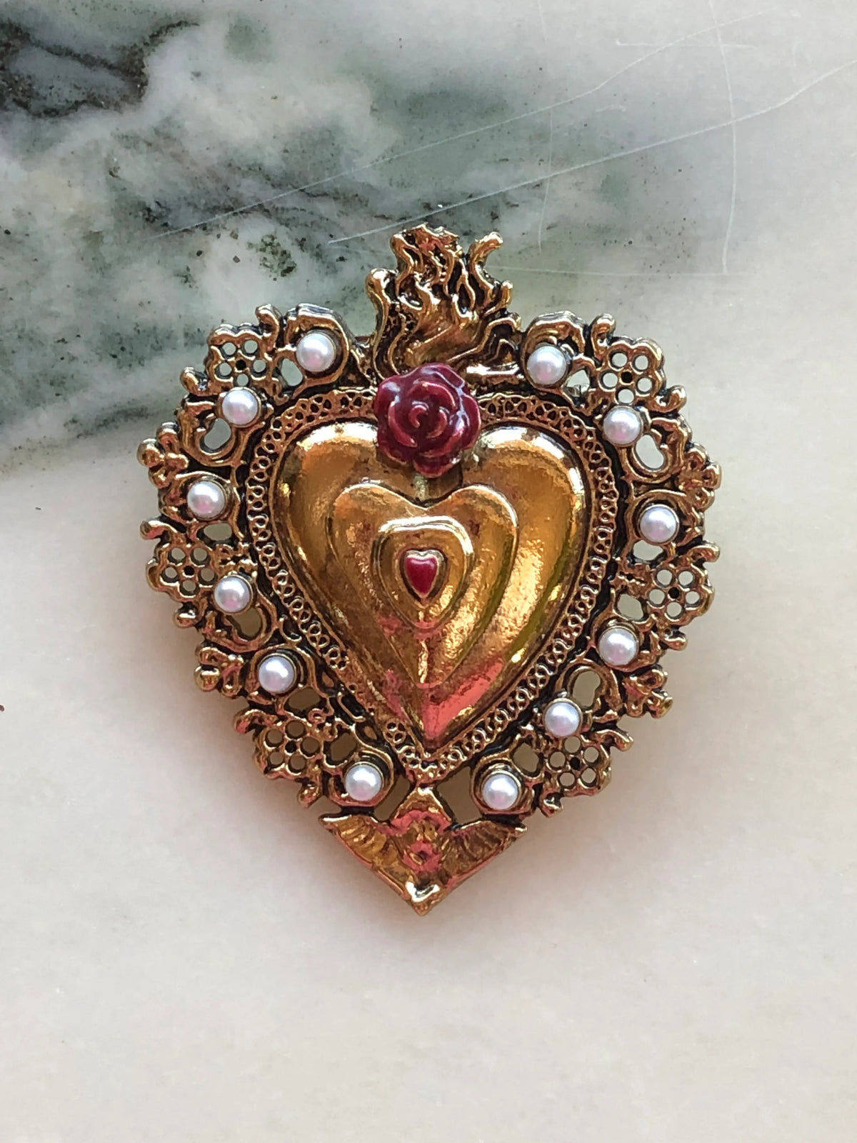 Broochella Men's Sacro Cuore Benedetta Heart Brooch in red, featuring an ornate gold-tone sacred heart with red rose detail, pearl embellishments and flame motif, photographed on a white marble background