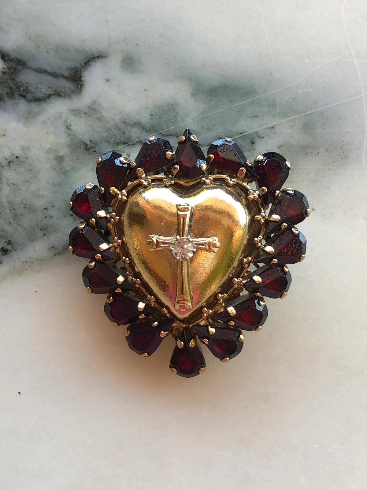 Broochella Men's Sacro Cuore Cristo Heart Brooch featuring a gold-tone sacred heart with cross and crystal centre, surrounded by deep garnet red faceted stones, photographed on a white marble background.