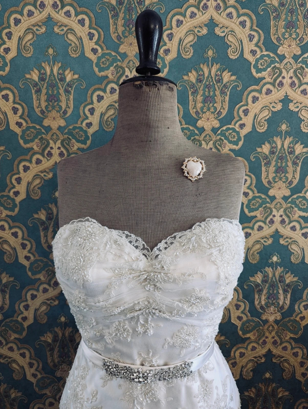 Broochella sacro cuore fiorentina white bridal heart shaped enamel brooch with gold metal base. Pictured on a mannequin that is wearing a white, beaded strapless wedding dress. The background is green and gold damask wallpaper. 