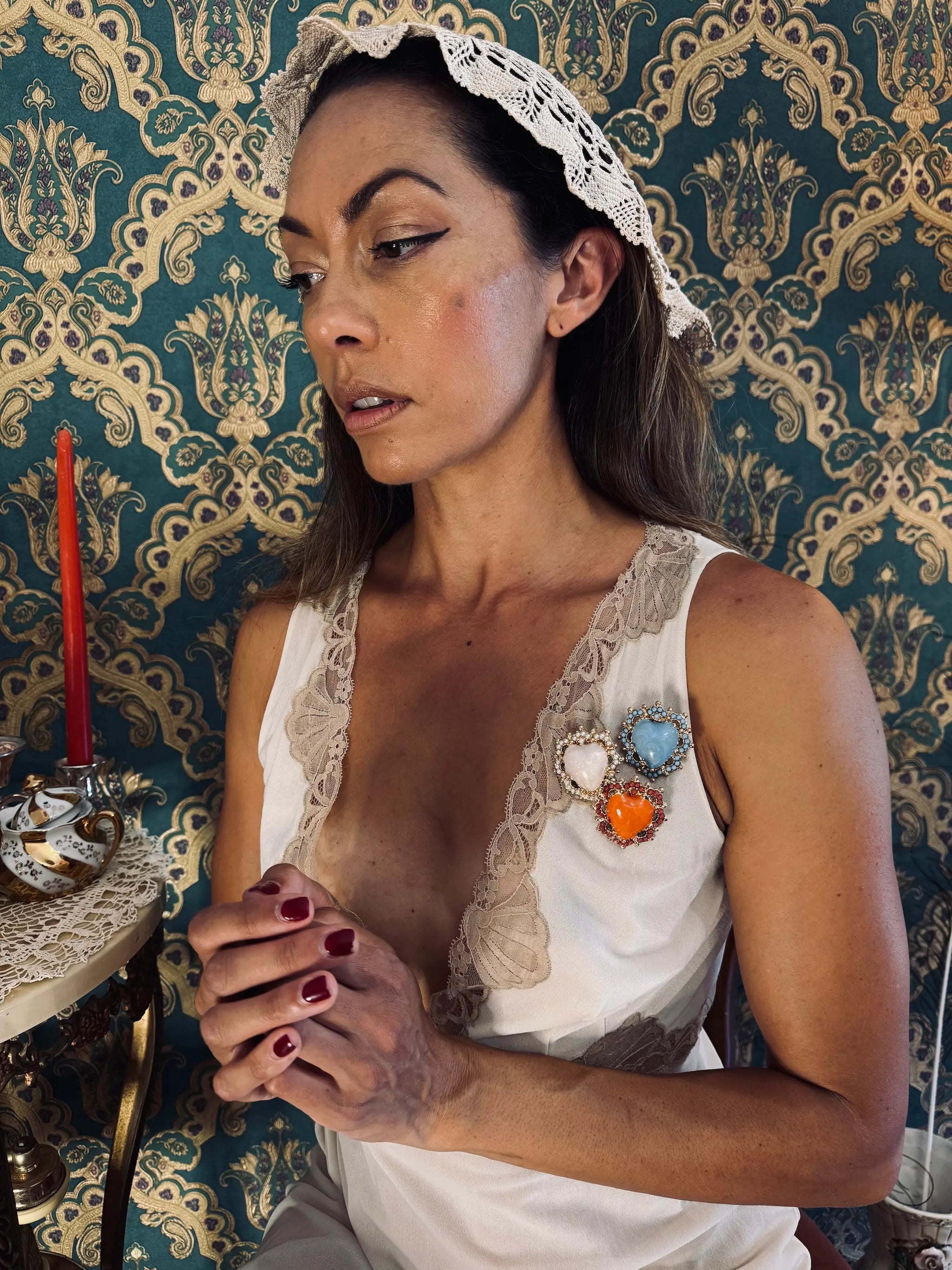 Broochella sacro cuore fiorentina brooches are pinned on a model in an ivory slip dress. She has dark hair and is wearing a veil. The background is green and gold damask wallpaper.