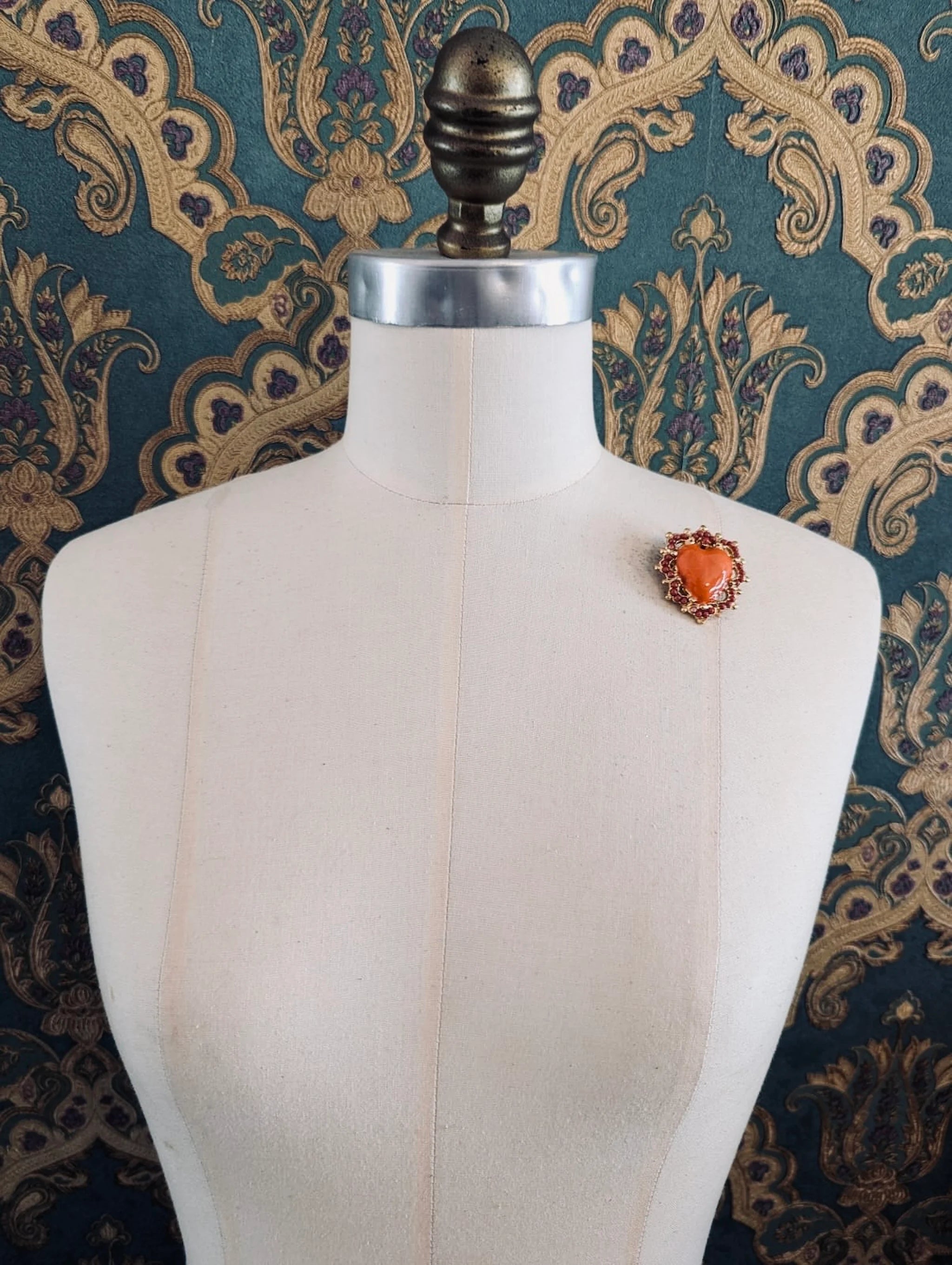 Broochella Sacro cuore fiorentina coral with gold metal base heart shaped brooch. Pinned to a mannequin. The background is green and gold damask wallpaper