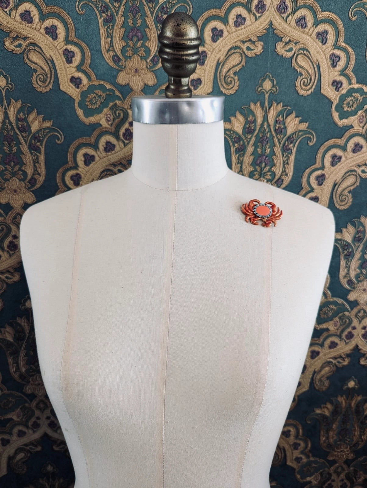 Broochella Sacro cuore granchio orange crab shaped brooch. Pinned to a mannequin. The background is green and gold damask wallpaper