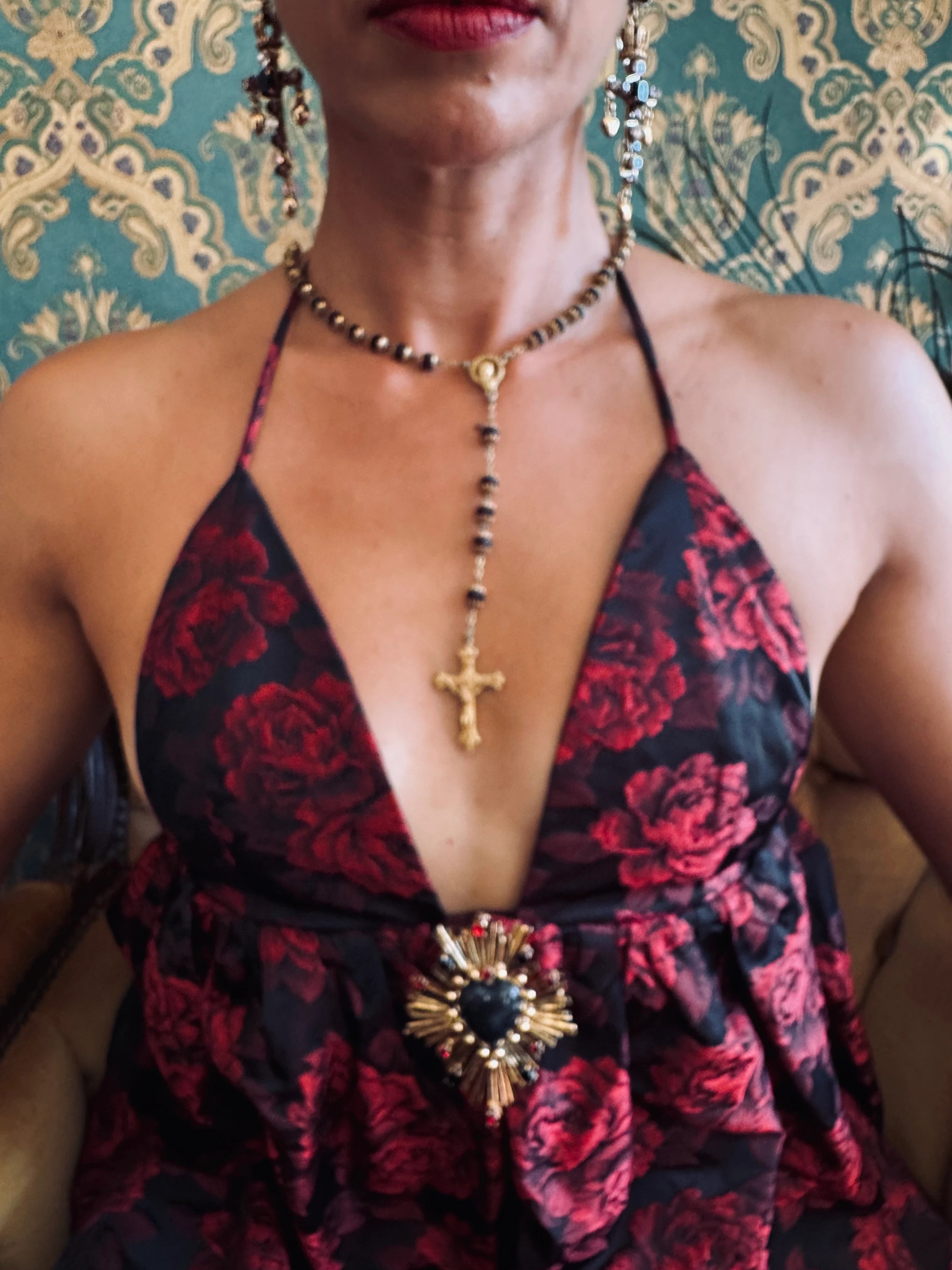 Broochella sacro cuore lucia brooch pinned on the dress of a model in a red and black dress. she is wearing a a crucifix necklace. The background is green and gold damask wallpaper