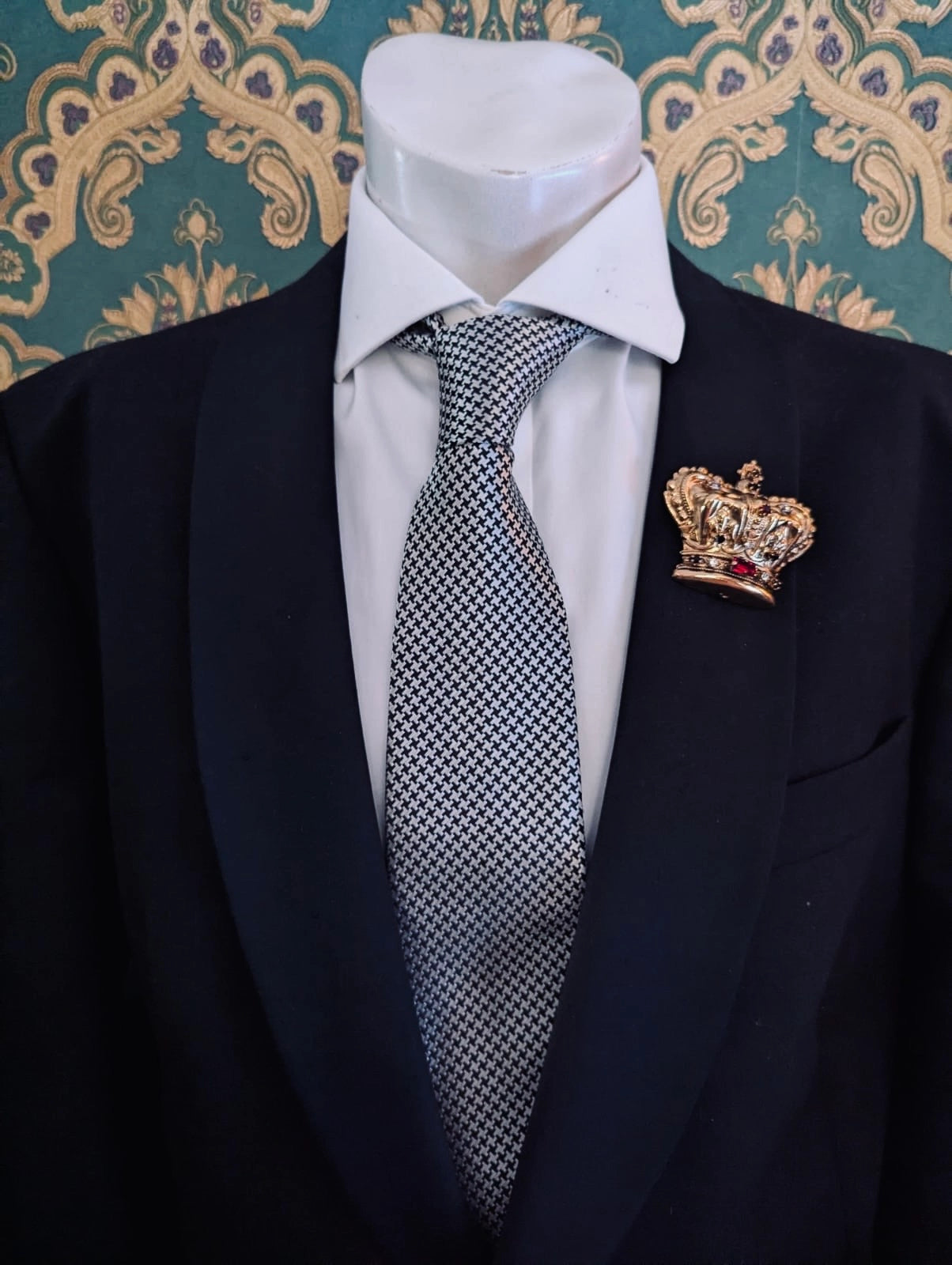Broochella sacro cuore men's regina gold metal crown brooch Pinned on a men's mannequin wearing a black suit and a houndstooth tie. The background is green and gold damask wallpaper. 