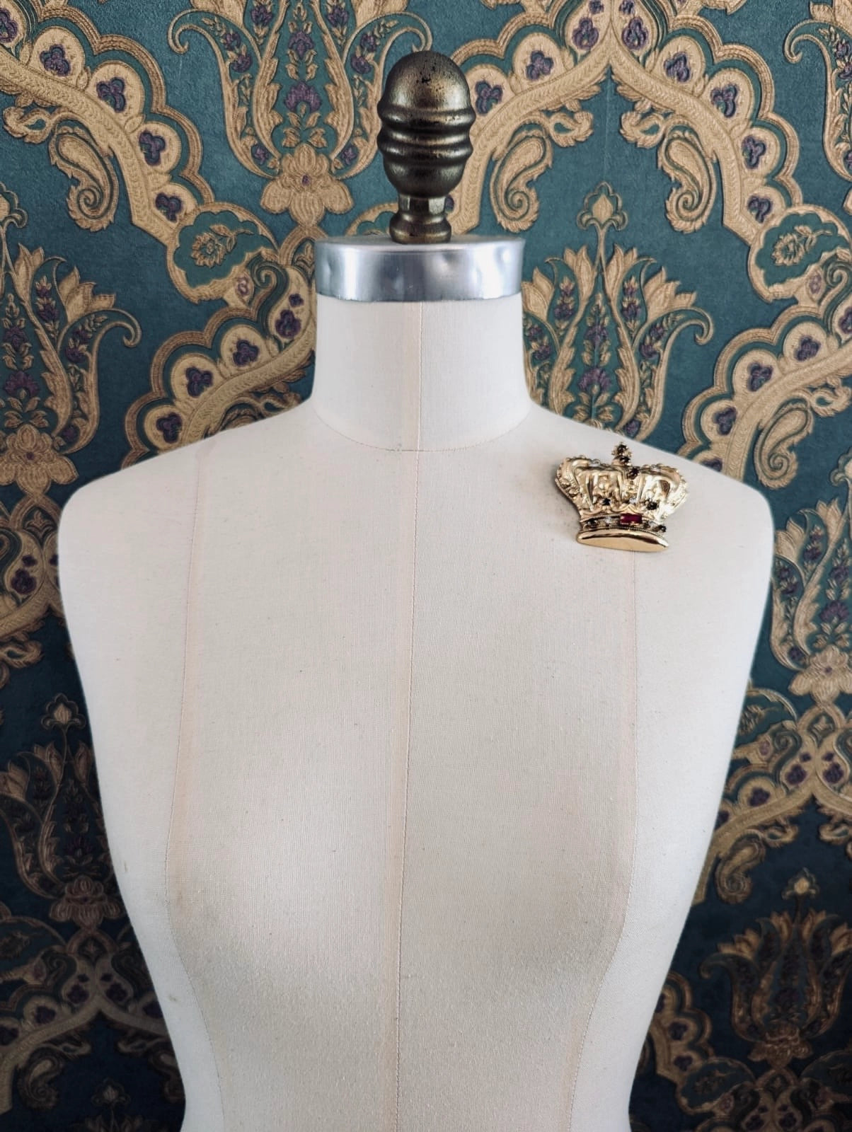 Broochella Sacro cuore regina gold designer crown brooch. Pinned to a mannequin. The background is green and gold damask wallpaper