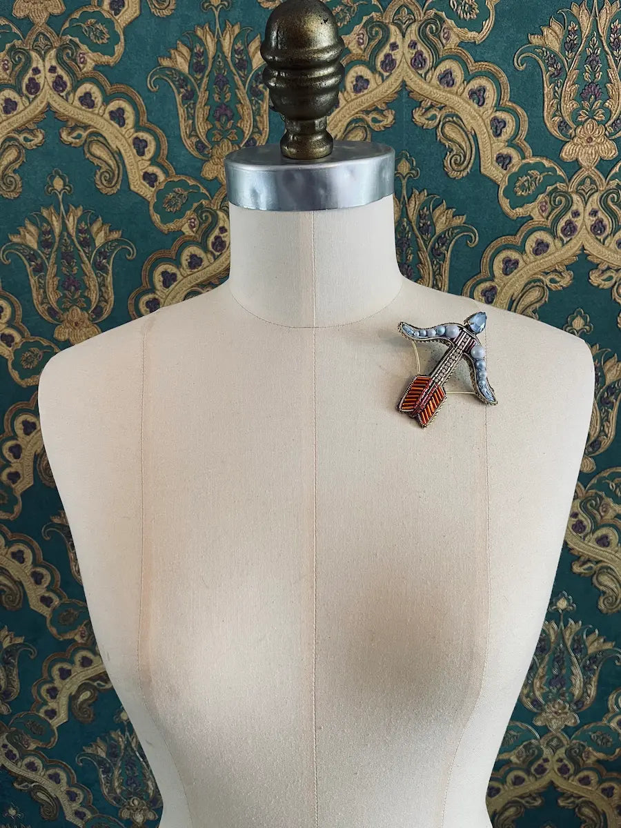 Broochella sagittarius hand beaded blue orange designer brooch. Styled on a mannequin. The background is green and gold damask wallpaper