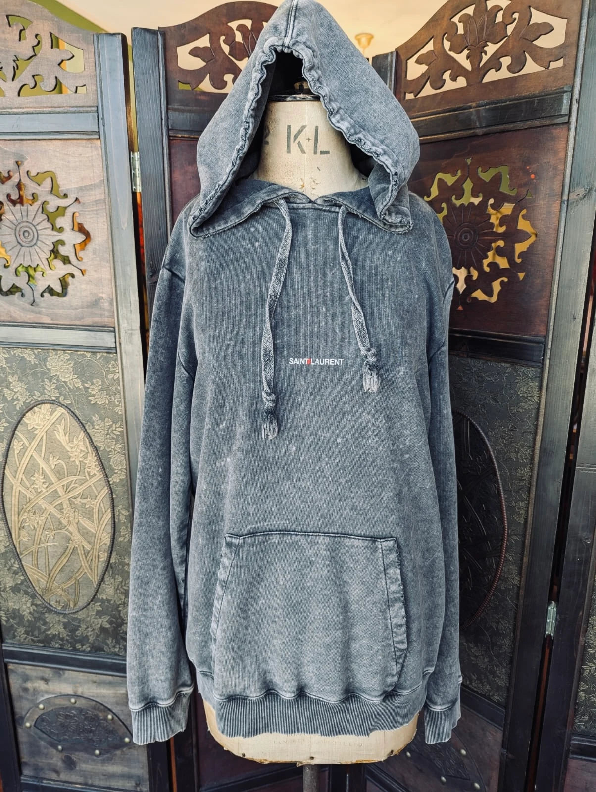 Broochella authentic designer Saint Laurent hoodie in distresses black. It is pictured on a mannequin. The background is a dark wood room divider.