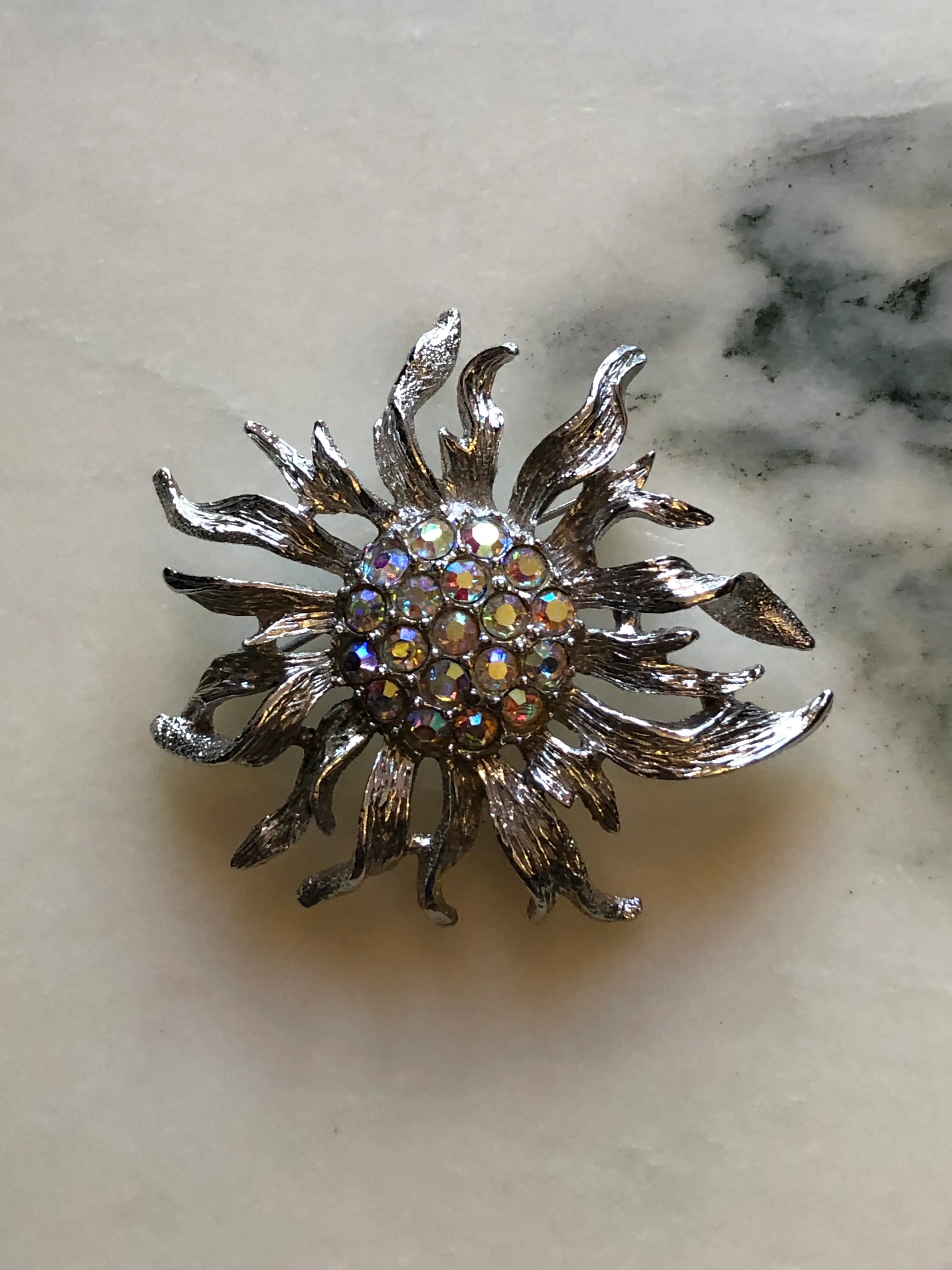 A silver-toned Sarah Coventry sunburst brooch featuring a central cluster of iridescent rhinestones, displayed on a light beige marble surface.