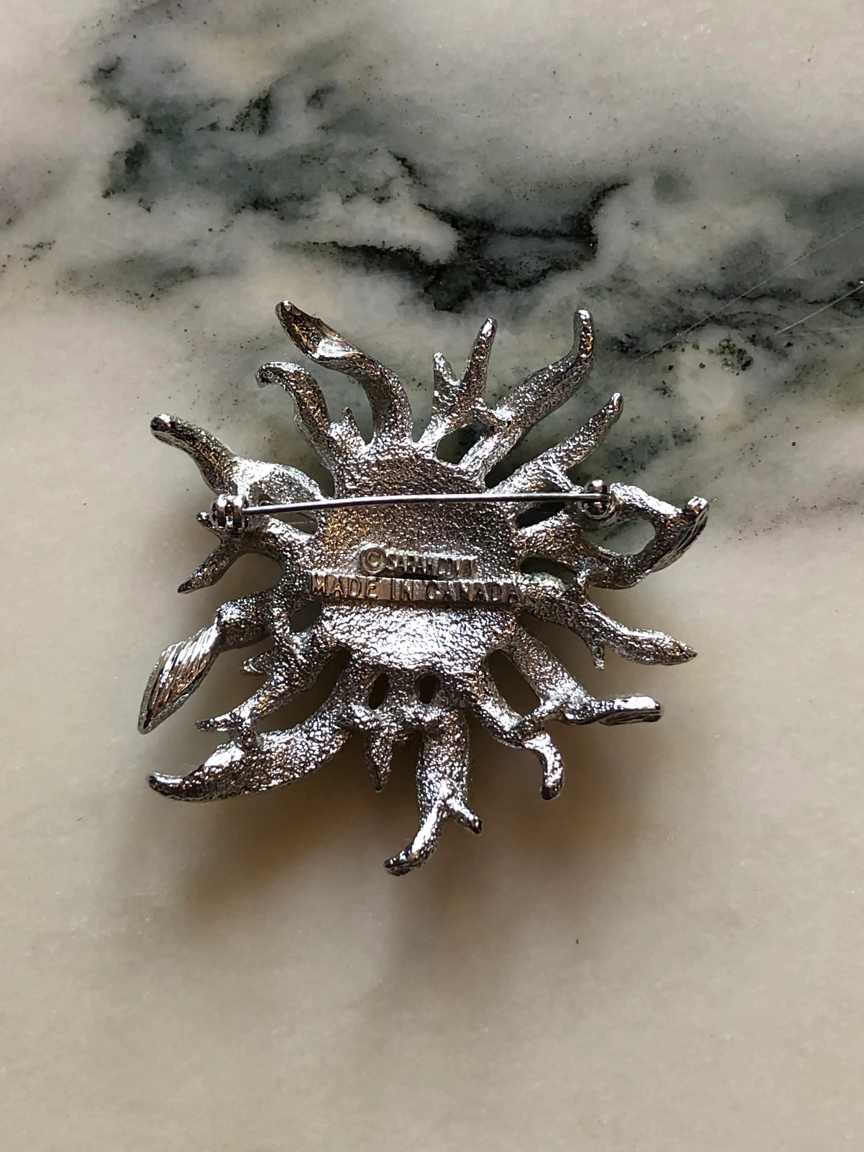 The back of a Broochella silver-toned Sarah Coventry sunburst brooch featuring a central cluster of iridescent rhinestones, displayed on a light beige marble surface.