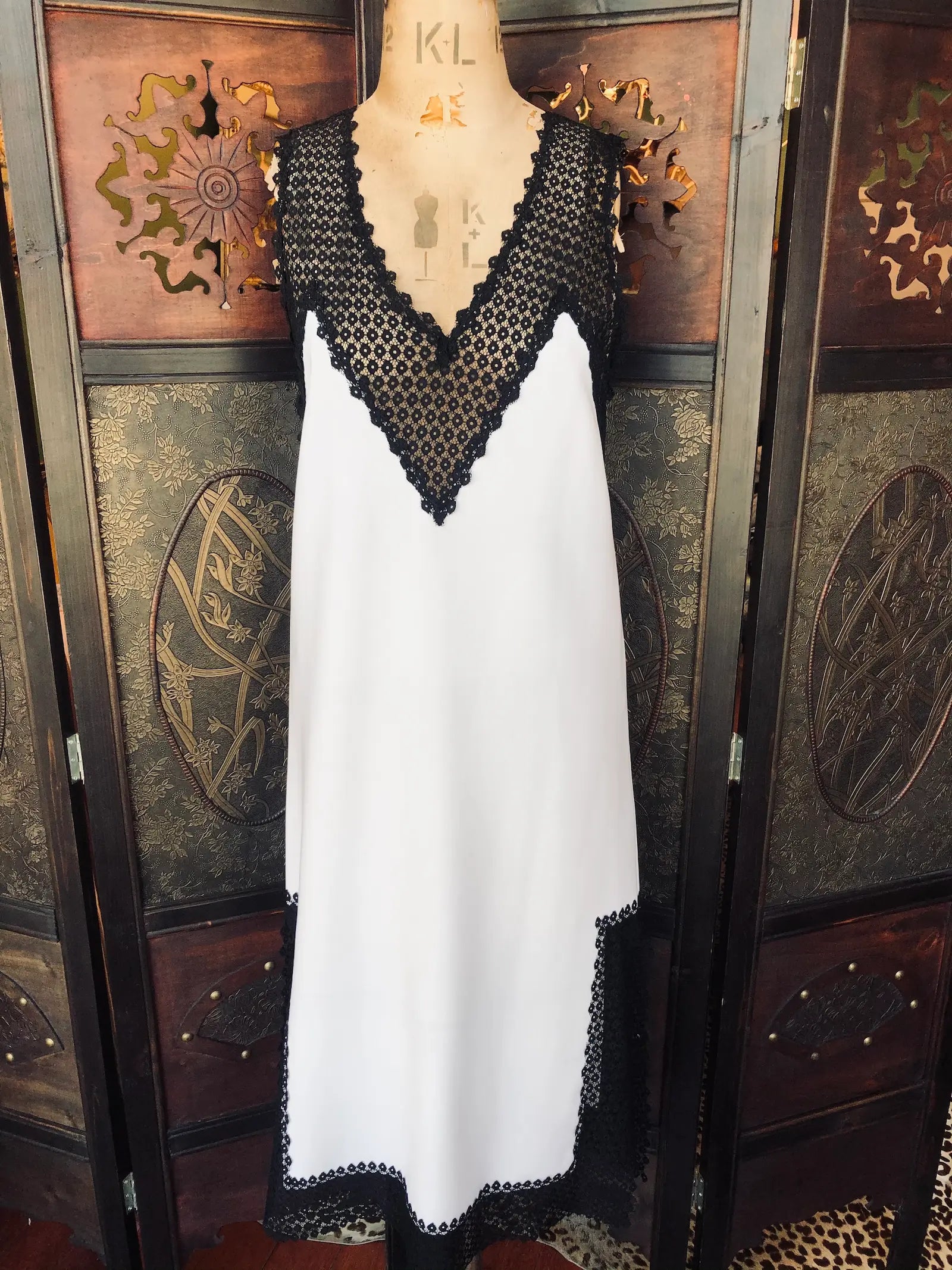 Broochella Scanlan Theodore midi slip dress. Size AU 6 It is Ivory with black lace detailing. It has Deep V lace neckline, 
sleeveless design, black lace-trimmed hem and a slip-style silhouette. It is pictured on a vintage mannequin. The background is a an ornate, dark wood, vintage room divider. 