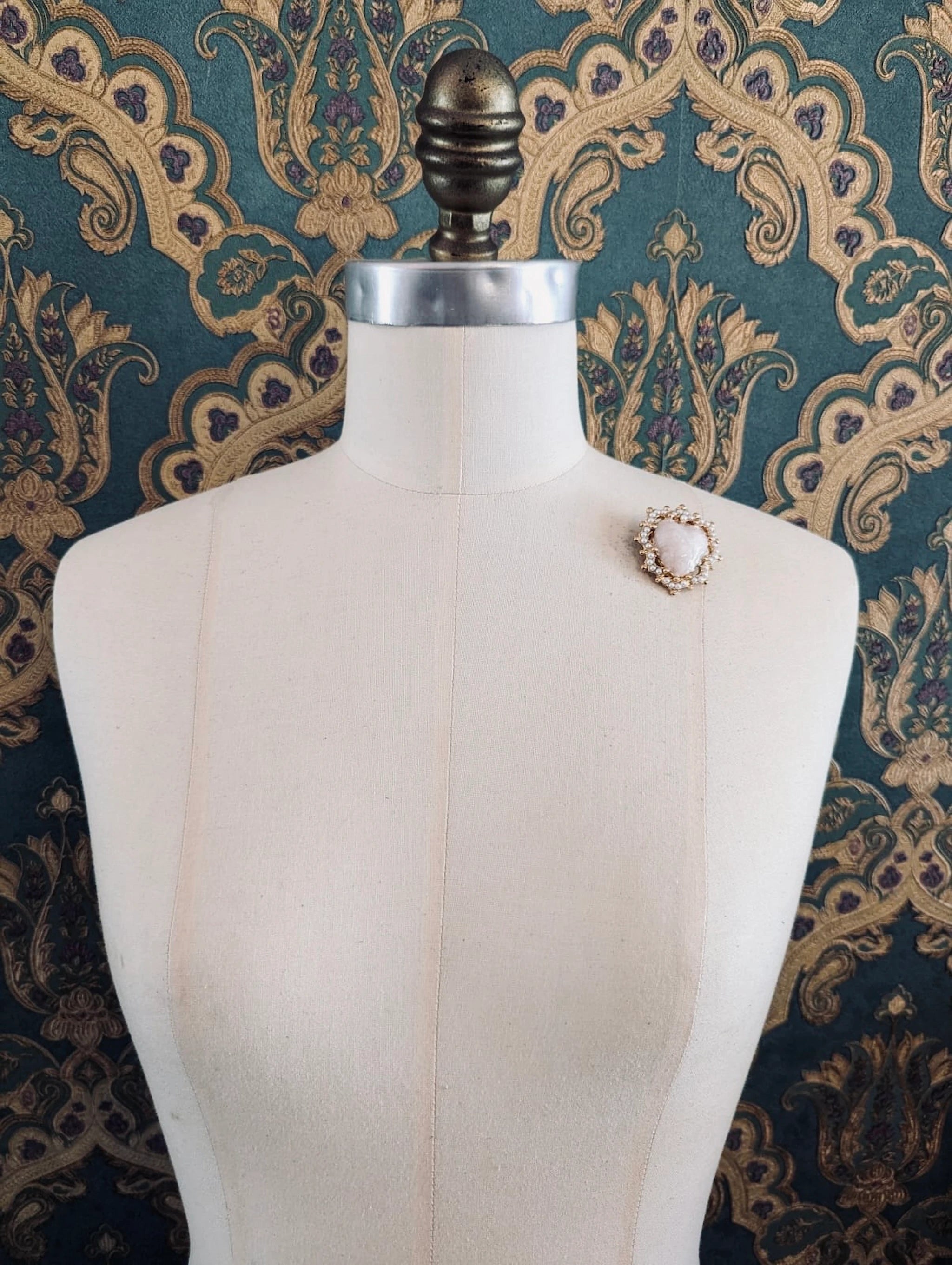 Broochella Sacro cuore fiorentina ivory with gold metal base heart shaped brooch. Pinned to a mannequin. The background is green and gold damask wallpaper.
