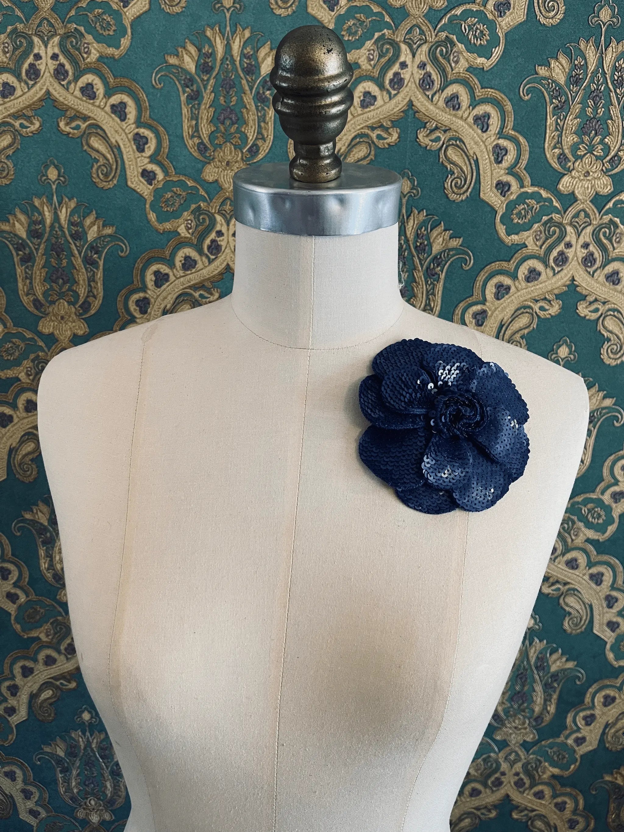 Broochella Scintillante handmade sequin flower brooch in blue. Pictured on a mannequin. The background is green and gold damask wallpaper