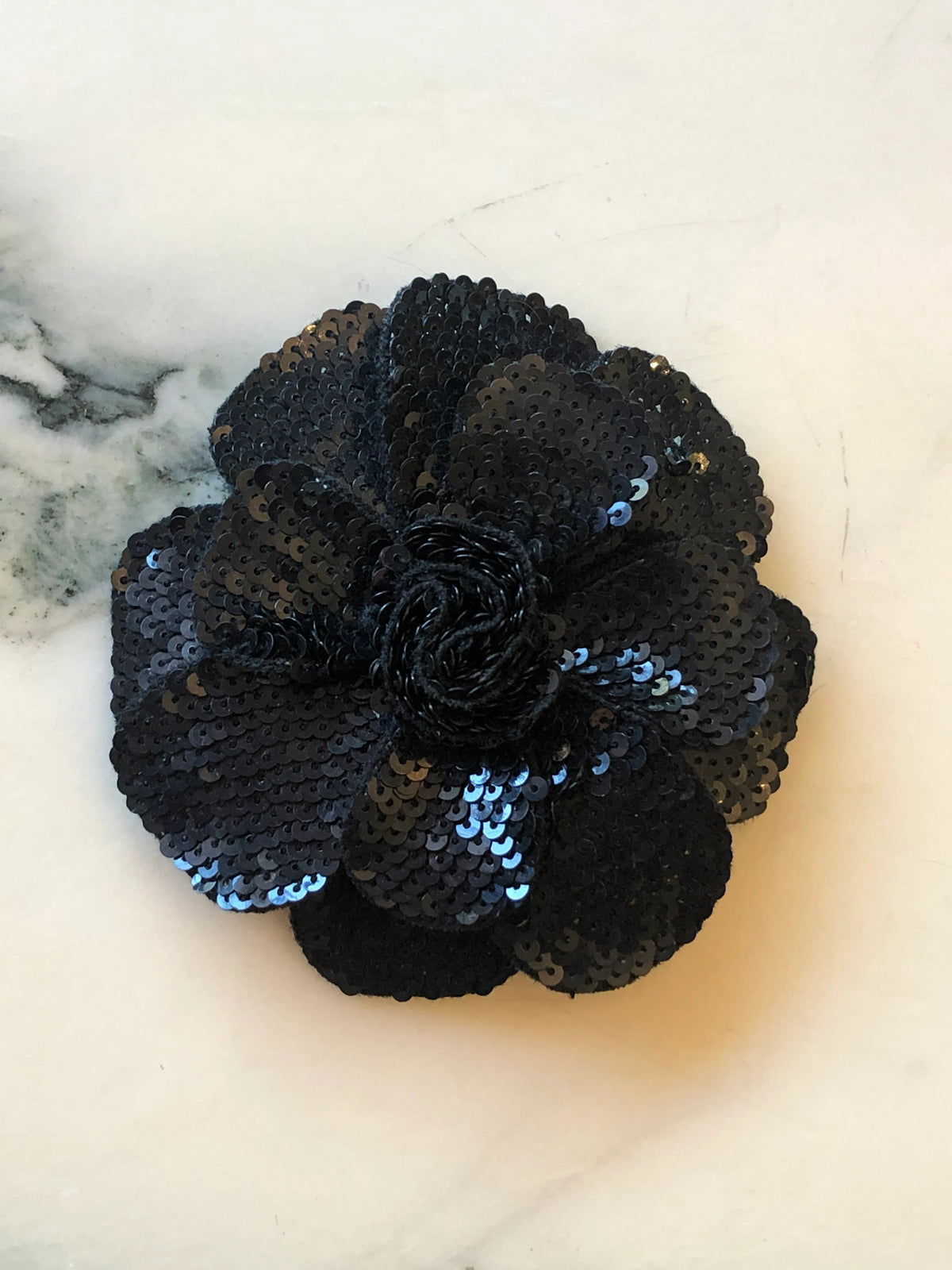 Broochella Scintillante Flower Brooch Pin in black, featuring layered sculptural petals embellished with tonal black sequins and a textured black centre on a cream marble background.
