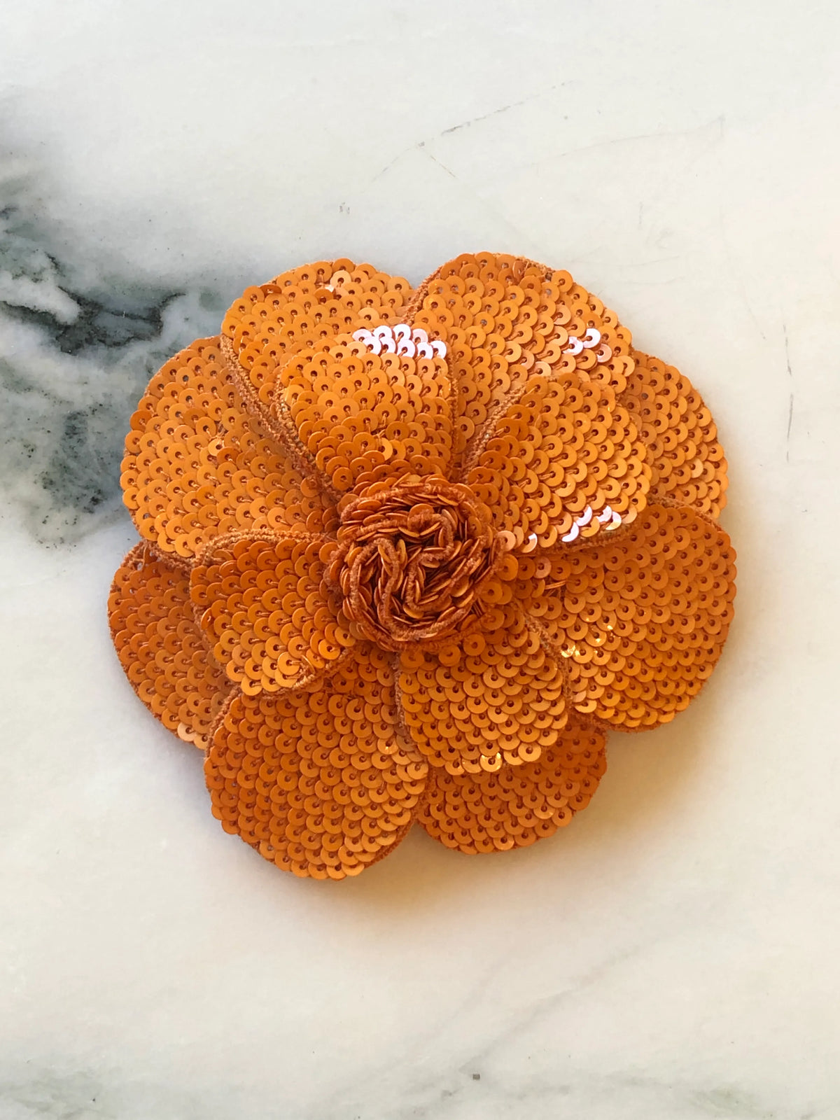 Broochella Scintillante Flower Brooch Pin in orange, featuring layered sculptural petals completely embellished with tonal orange sequins and a textured hand-finished centre on a cream marble background.