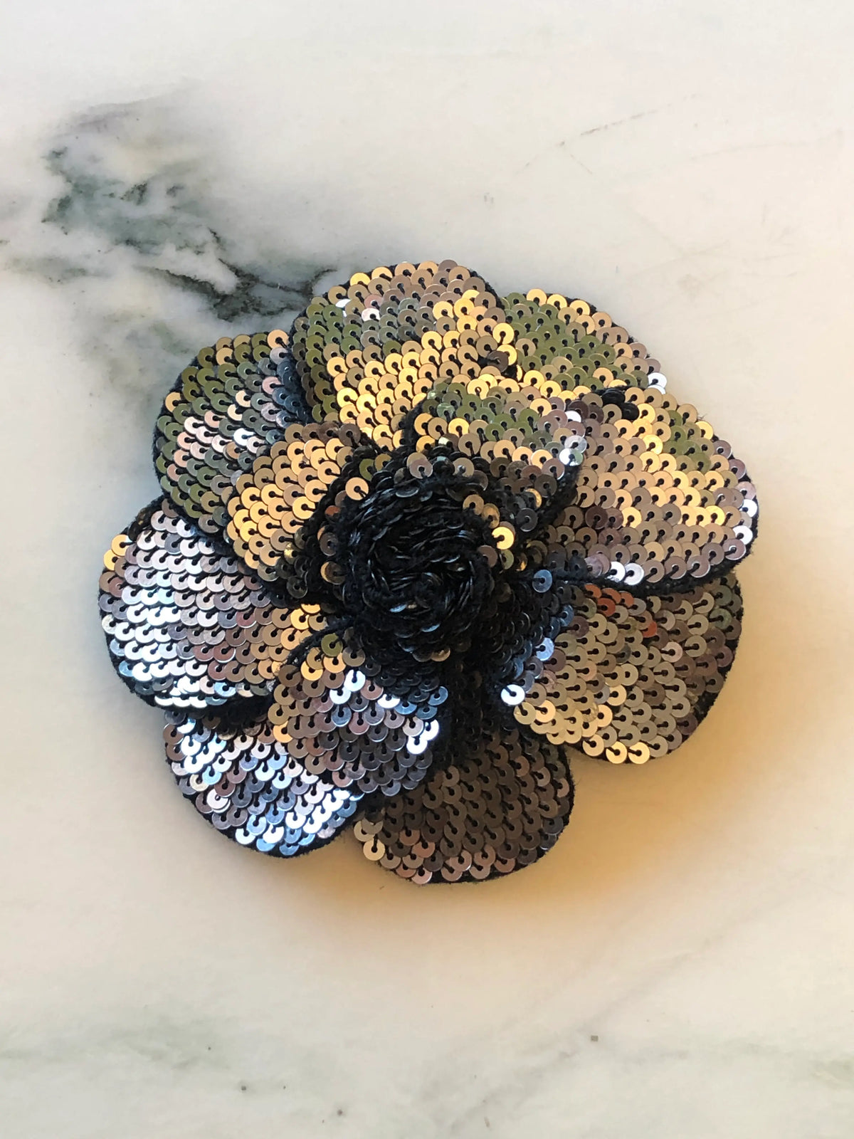 Broochella Scintillante Flower Brooch Pin in silver, featuring layered sculptural petals embellished with shimmering silver, pewter and gold-toned sequins with a black textured centre on a cream marble background.