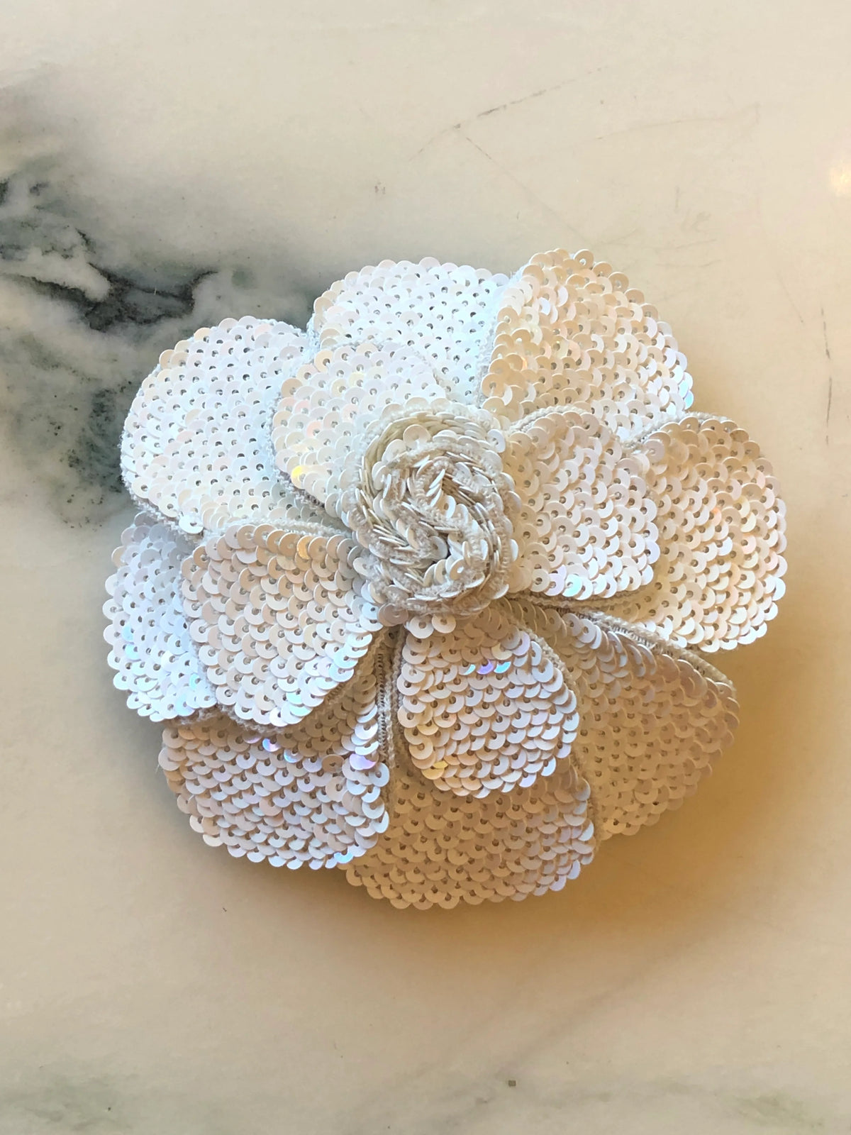 Broochella Scintillante Flower Brooch Pin in white, featuring layered sculptural petals completely embellished with luminous white sequins and a textured white centre on a cream marble background.