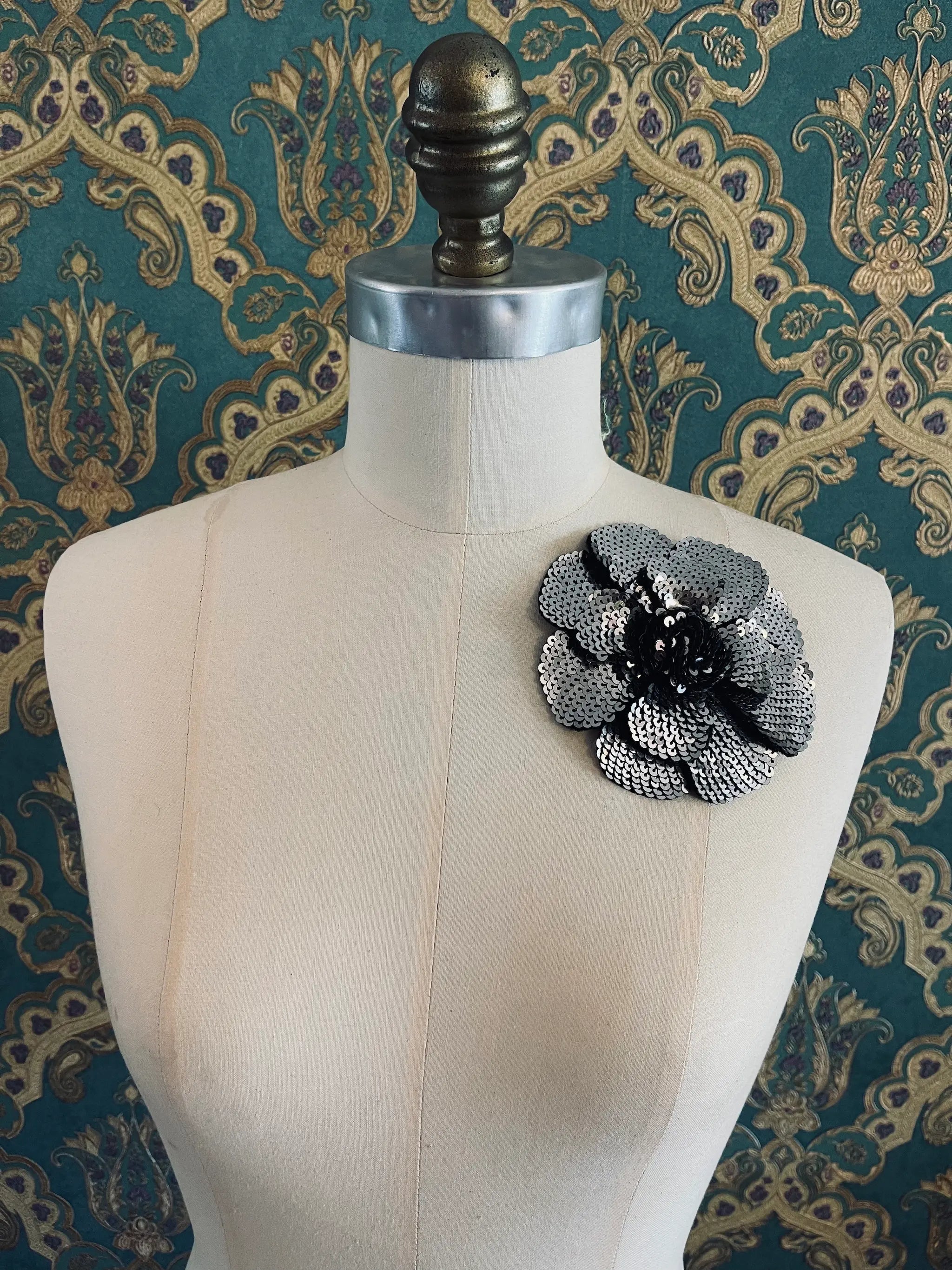 Broochella Scintillante handmade sequin flower brooch in silver. Pictured on a mannequin. The background is green and gold damask wallpaper