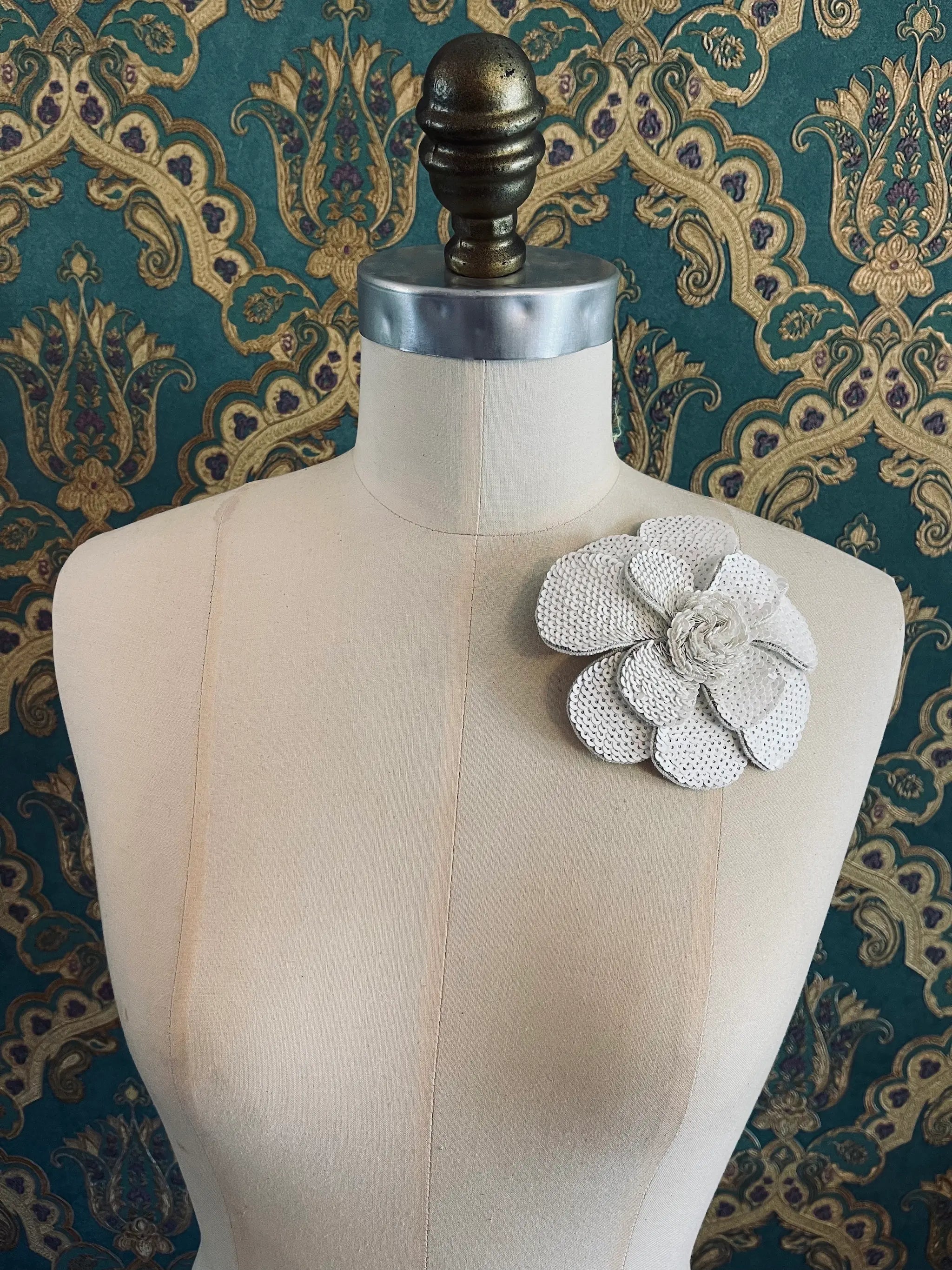 Broochella Scintillante handmade sequin flower brooch in white. Pictured on a mannequin. The background is green and gold damask wallpaper