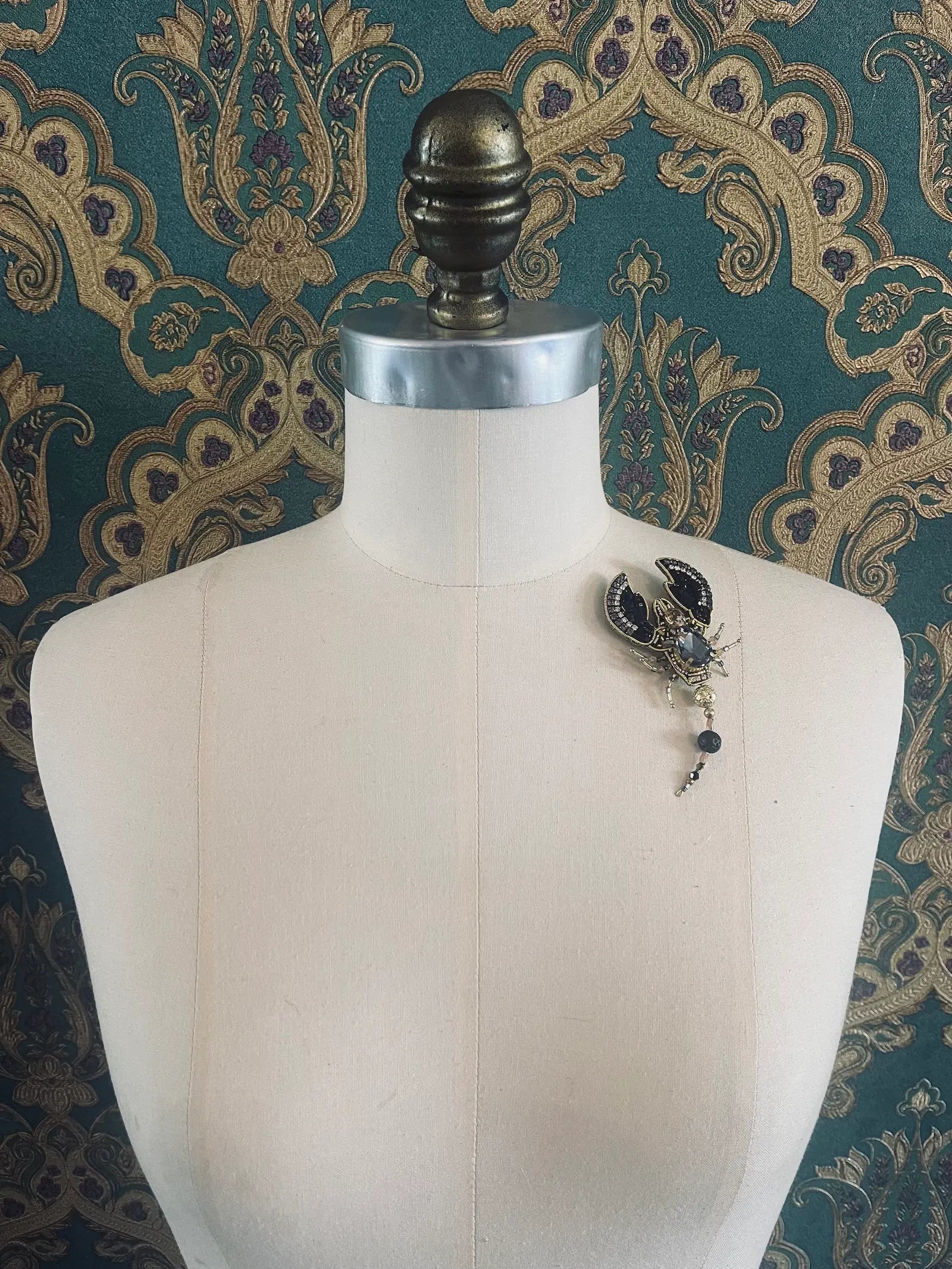 Broochella hand beaded black and gold scorpion brooch. Pictured on a mannequin. The background is green and gold damask wallpaper