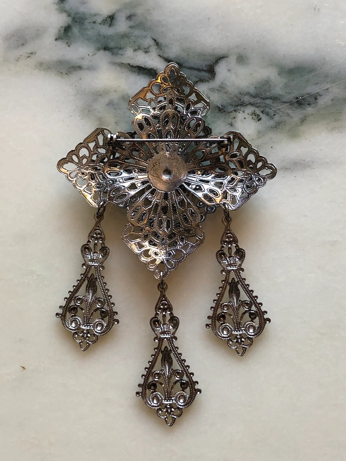 The back view of Broochella Serafina Vintage Silver Filigree Drop Brooch featuring ornate layered silver-tone filigree, a crystal floral centre and three decorative hanging drops on a cream and grey marble background.
