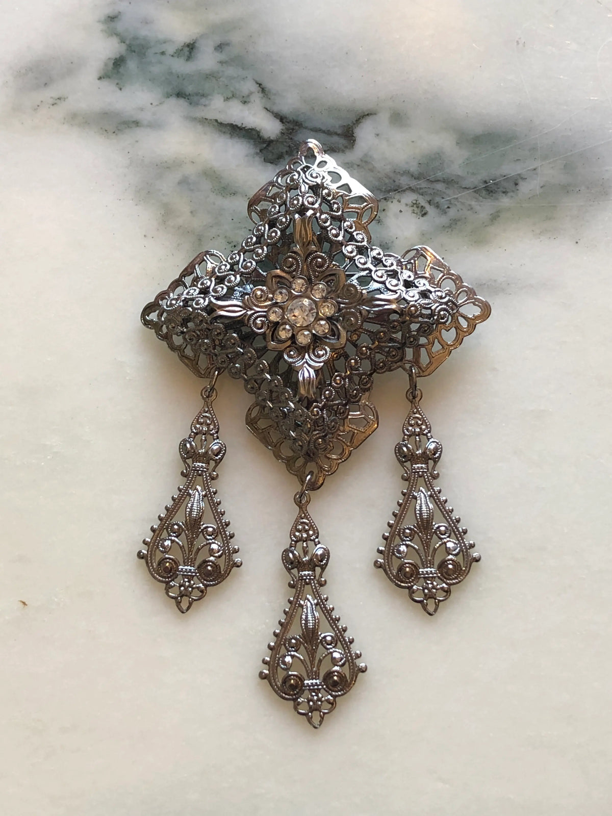Broochella Serafina Vintage Silver Filigree Drop Brooch featuring ornate layered silver-tone filigree, a crystal floral centre and three decorative hanging drops on a cream and grey marble background.