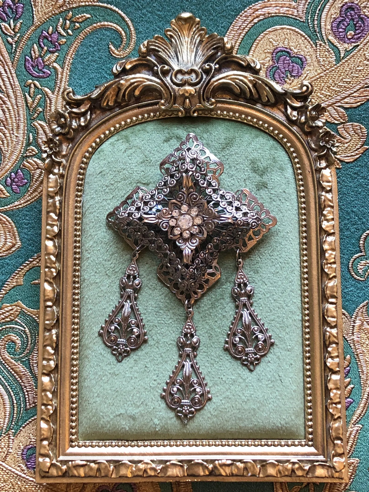 Broochella Serafina Vintage Silver Filigree Drop Brooch featuring ornate layered silver-tone filigree, a crystal floral centre and three decorative hanging drops. It is pictured on a green velvet tray with an ornate gold frame. The background is green and gold damask wallpaper