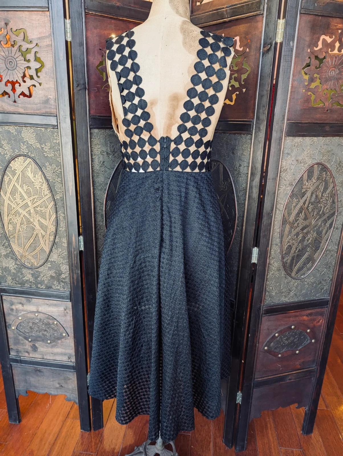 The back view of Broochella Shakuhachi Black Cut-Out Midi Dress – Size S. Pictured on a mannequin. The background is an ornate dark wood room divider. 