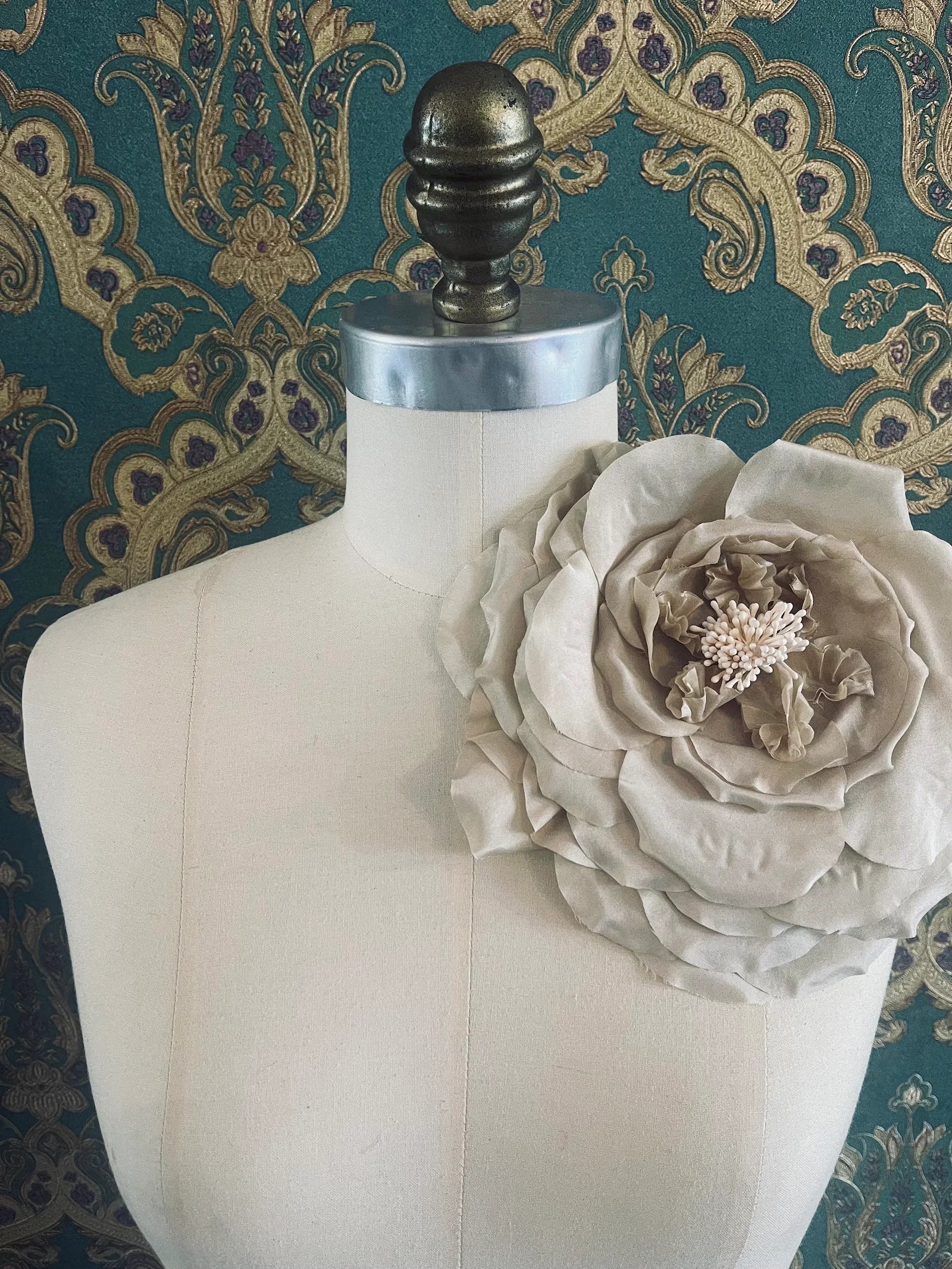 Broochella Signora Fiorita large handmade fabric flower brooch in beige. Broochella. Pictured on a mannequin. The background is green and gold damask wallpaper