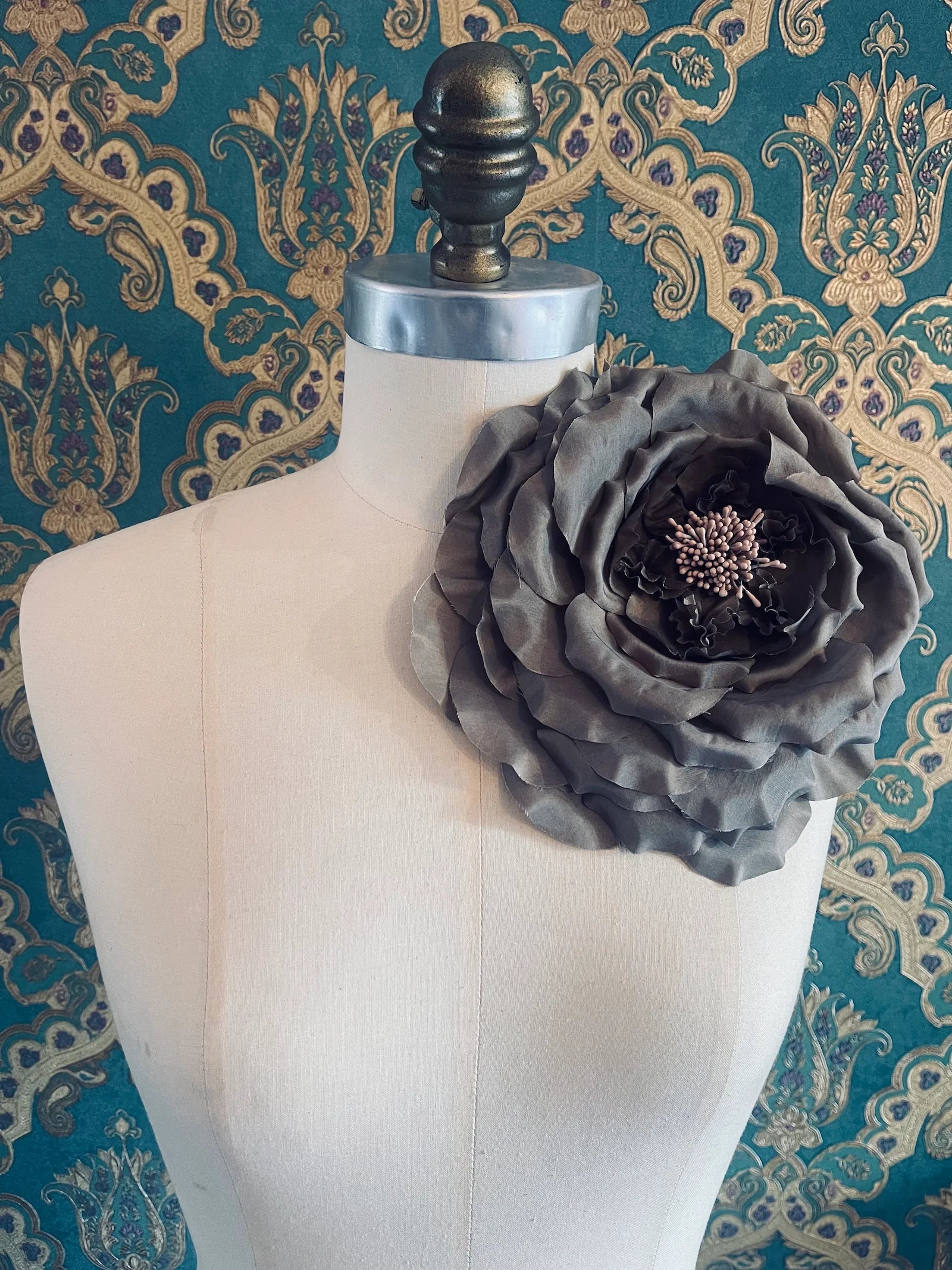 Broochella Signora Fiorita large handmade fabric flower brooch in brown. Broochella. Pictured on a mannequin. The background is green and gold damask wallpaper
