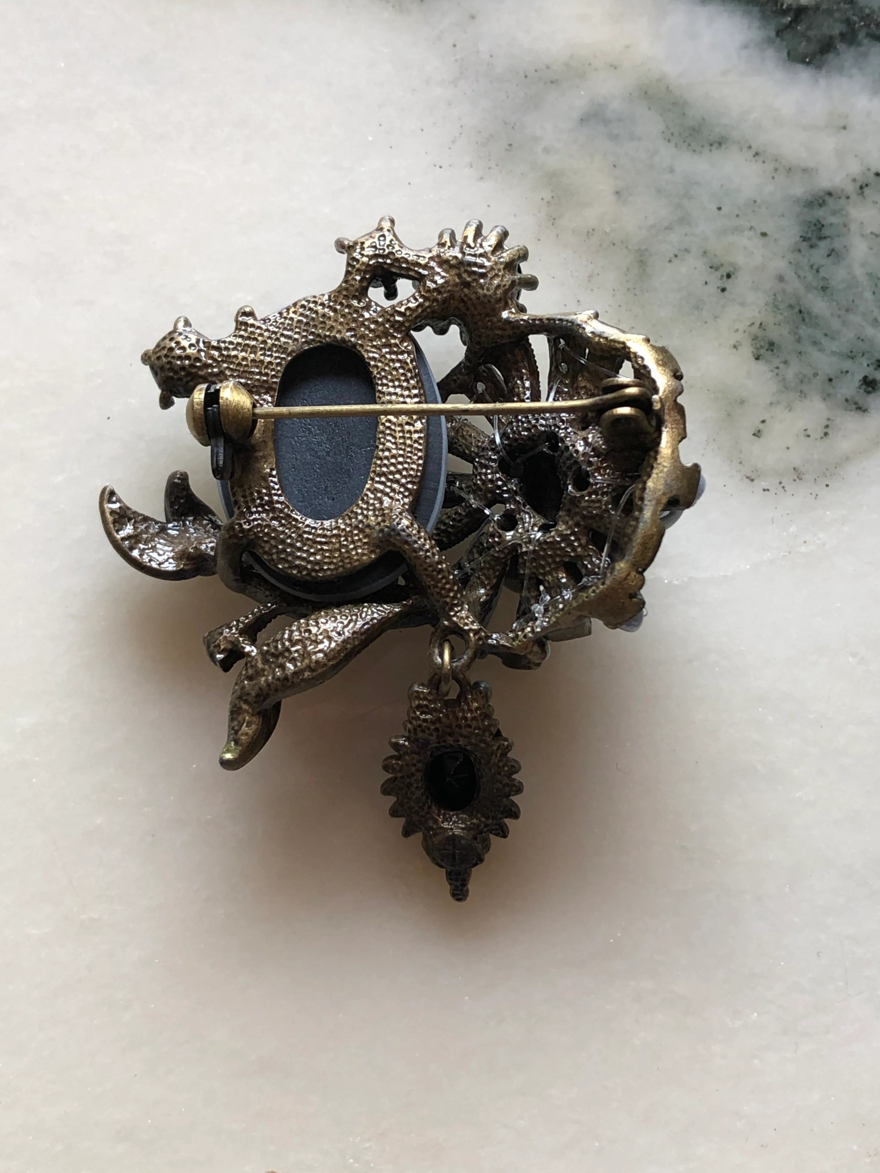 The back view of Broochella Signora Noir collectable brooch with a grey profile cameo, charcoal faux pearls, black crystals and antiqued gold-tone leaves. The background is white marble.