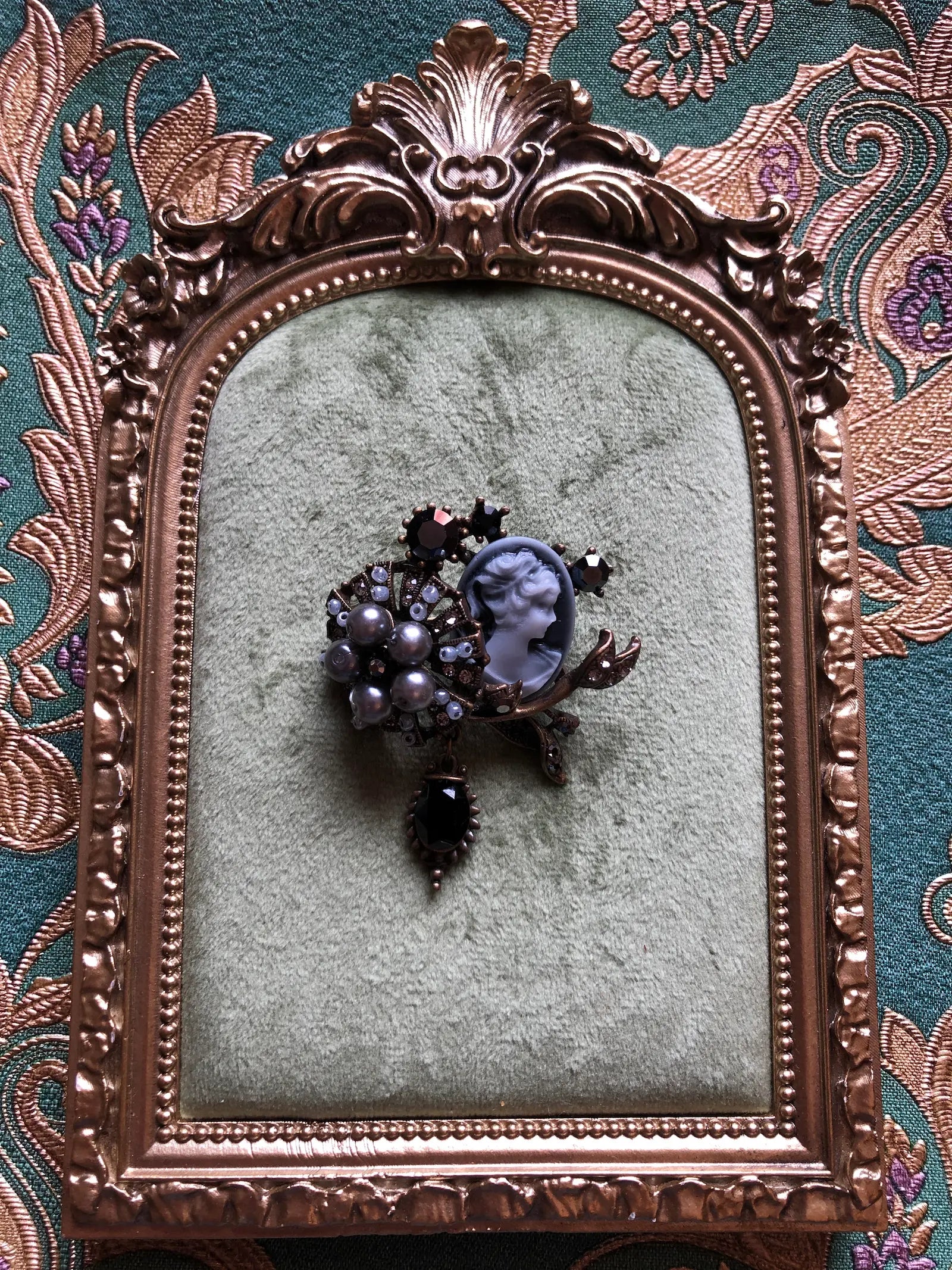 Broochella Signora Noir collectable brooch with a grey profile cameo, charcoal faux pearls, black crystals and antiqued gold-tone leaves. Pictured on a green velvet tray with an ornate gold frame. The background is green and gold damask wallpaper
