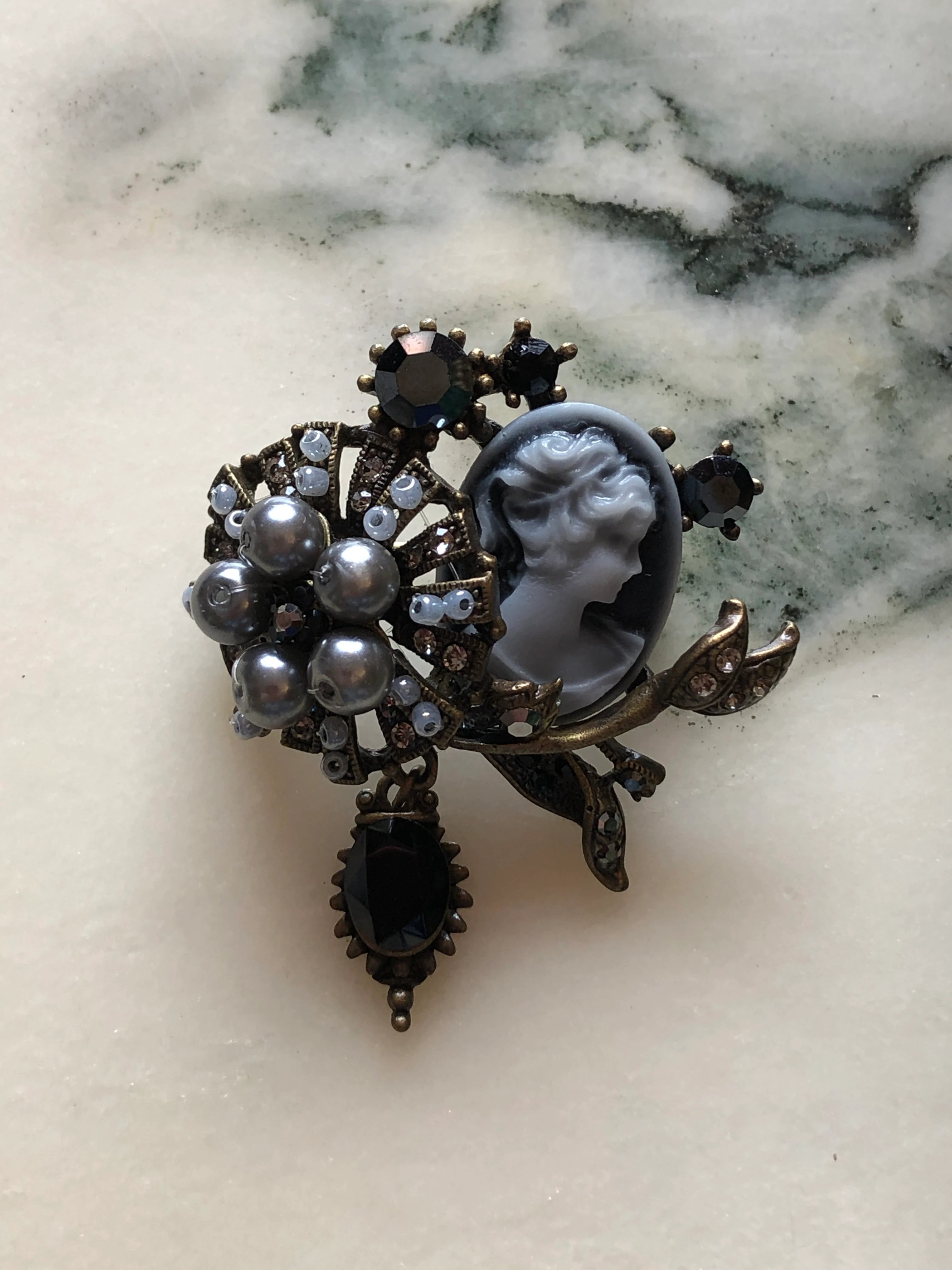 Broochella Signora Noir collectable brooch with a grey profile cameo, charcoal faux pearls, black crystals and antiqued gold-tone leaves. The background is white marble.