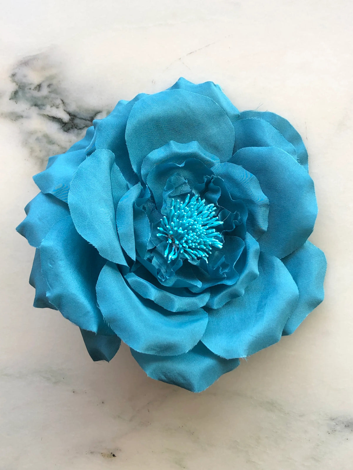 Broochella Men's Signorina Fioritina Flower Brooch Pin in blue with layered fabric petals and a matching stamen centre, photographed on a white marble background with grey veining.