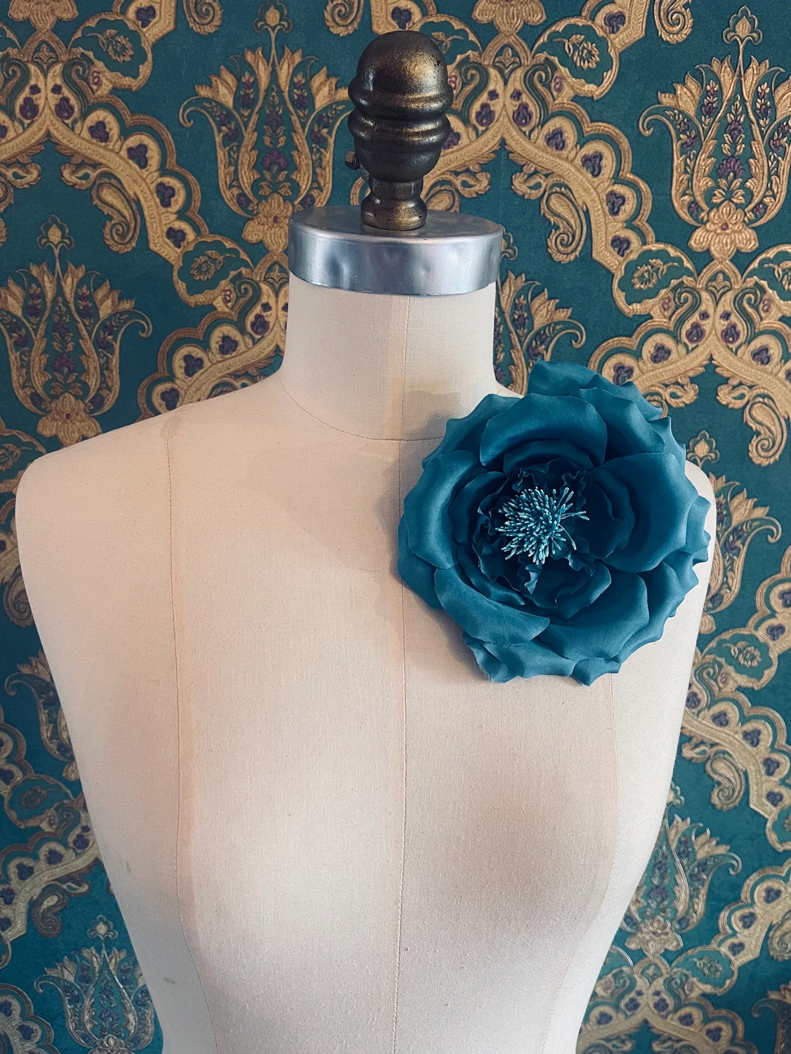 Broochella signorina fioritina handmade fabric flower brooch in blue. Pictured on a mannequin. The background is green and gold damask wallpaper